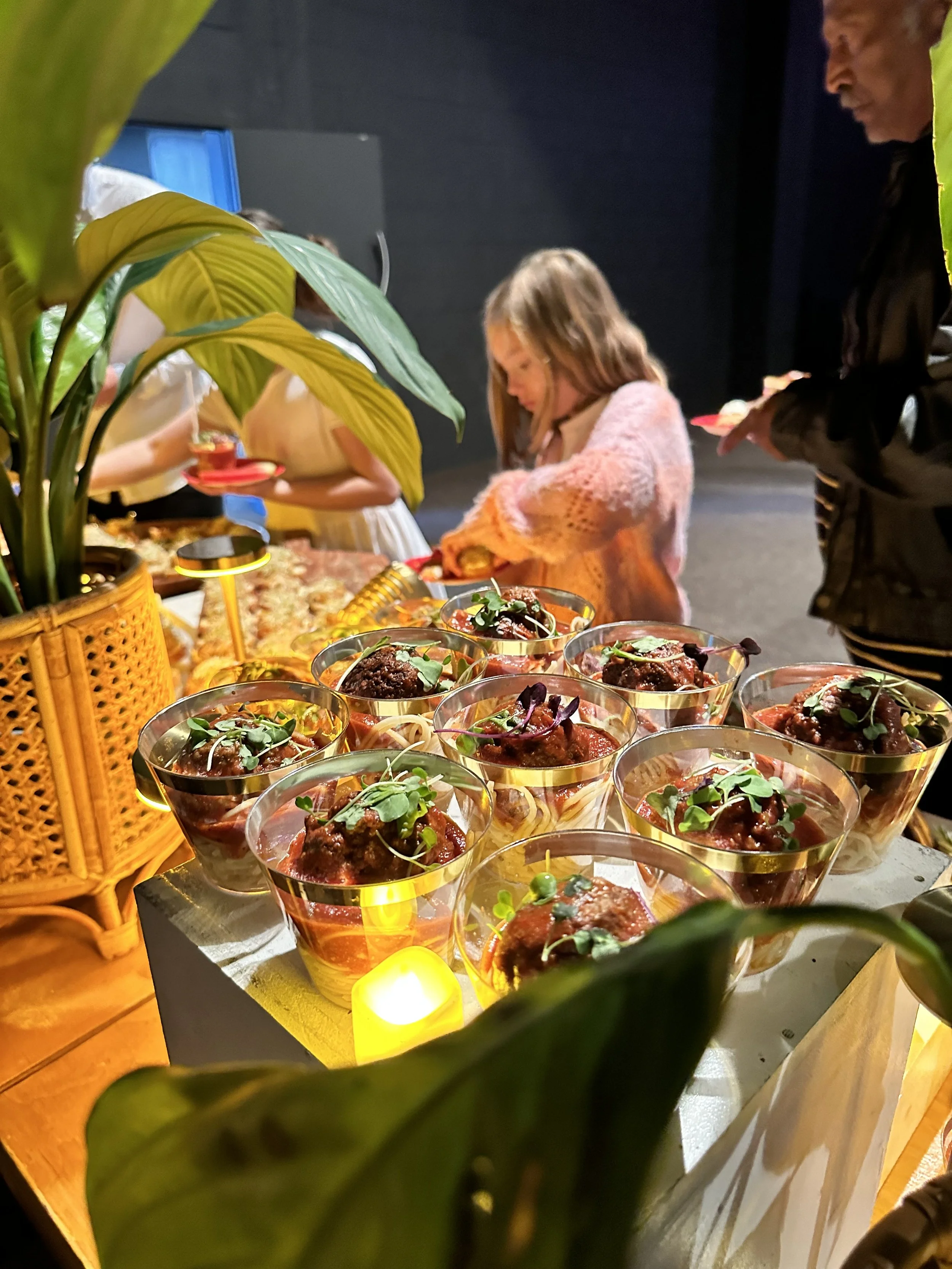 A dessert table with small glasses filled with a layered dessert topped with microgreens, surrounded by green leaves and a candle, with children and an adult in the background.
