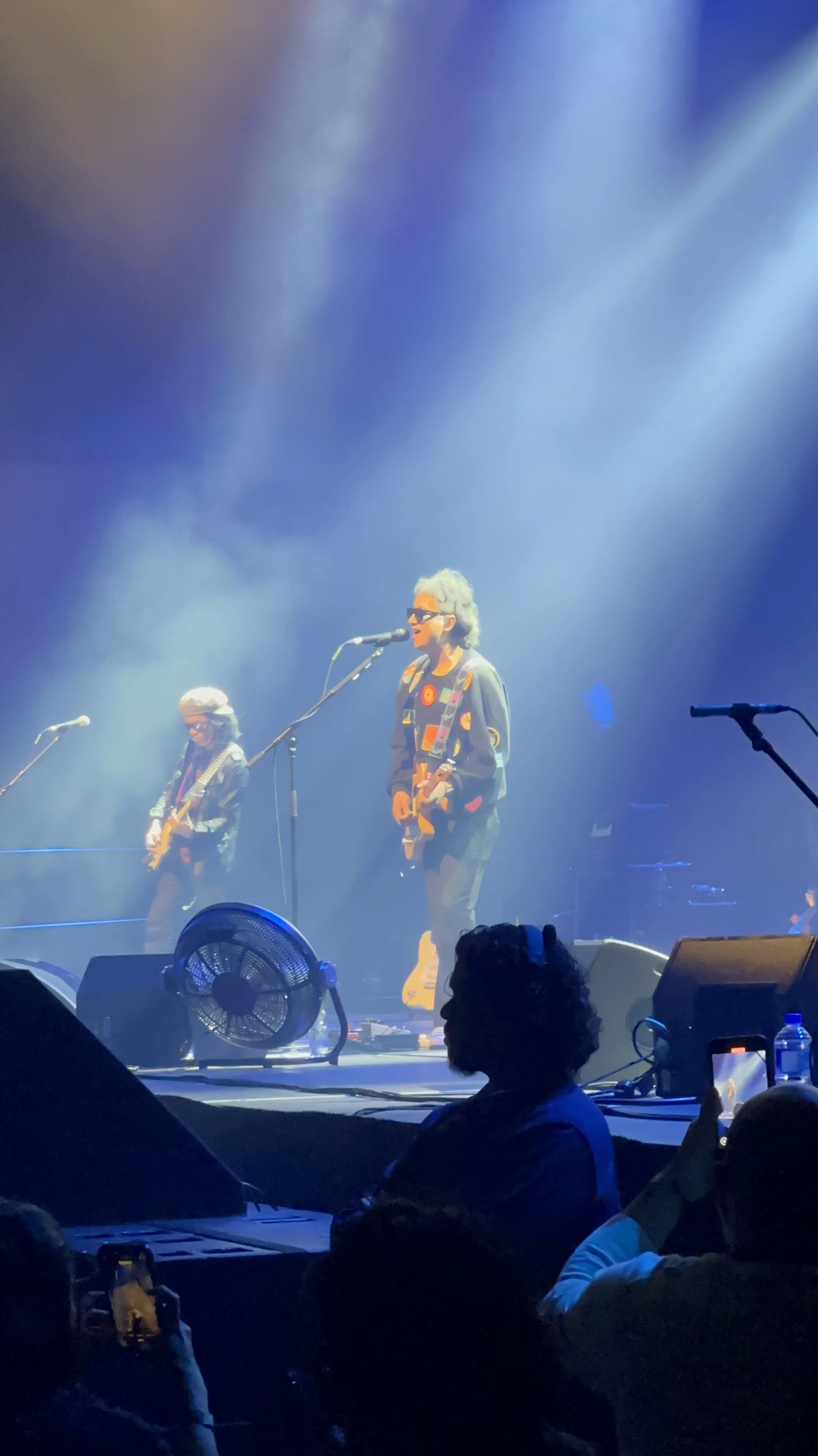 Musicians performing on stage with blue lighting and fog, with one person singing into a microphone while playing guitar and others playing instruments.