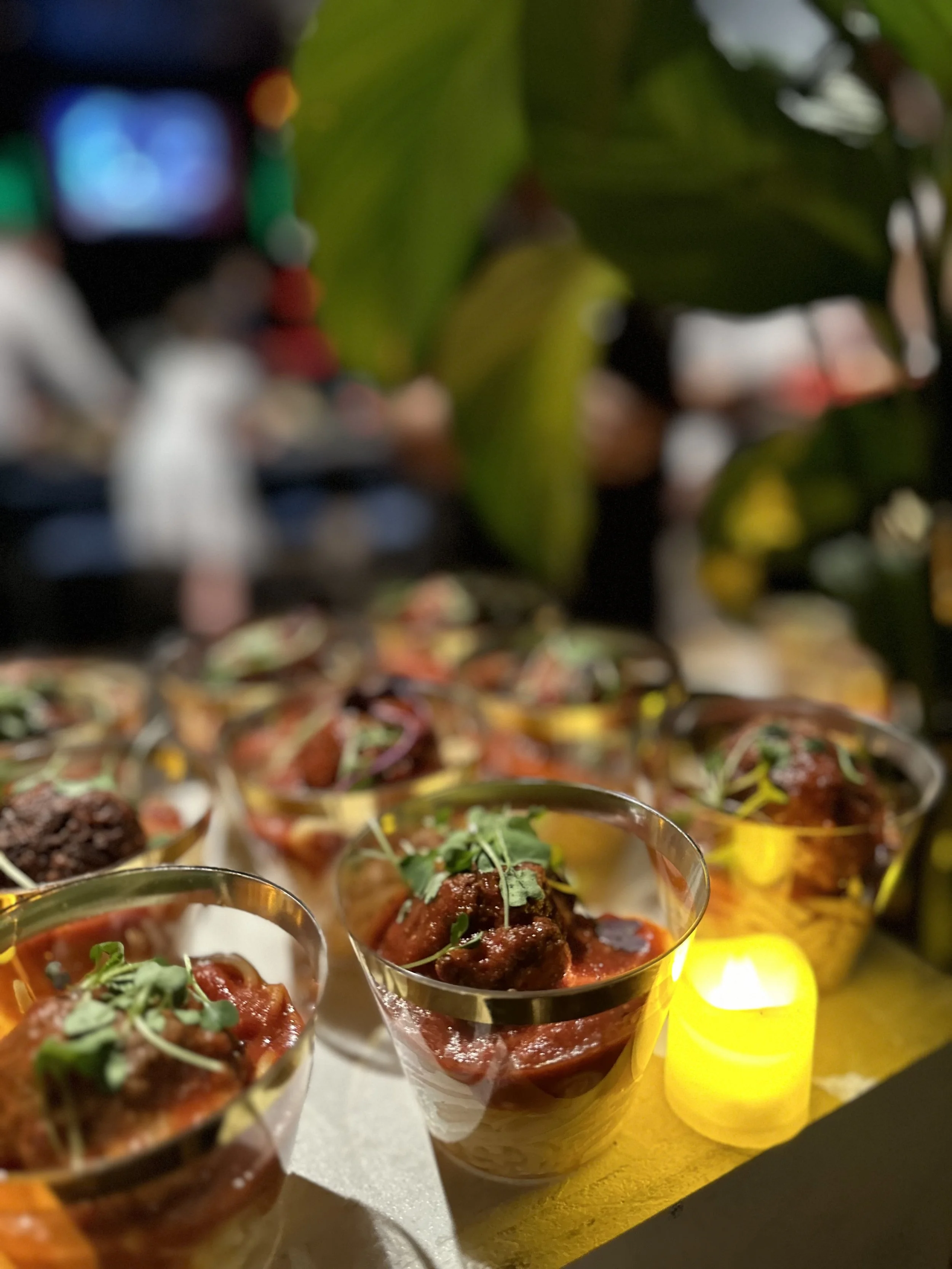 Glass cups filled with meatball dishes garnished with microgreens, set on a yellow surface with greenery and a candle beside them.