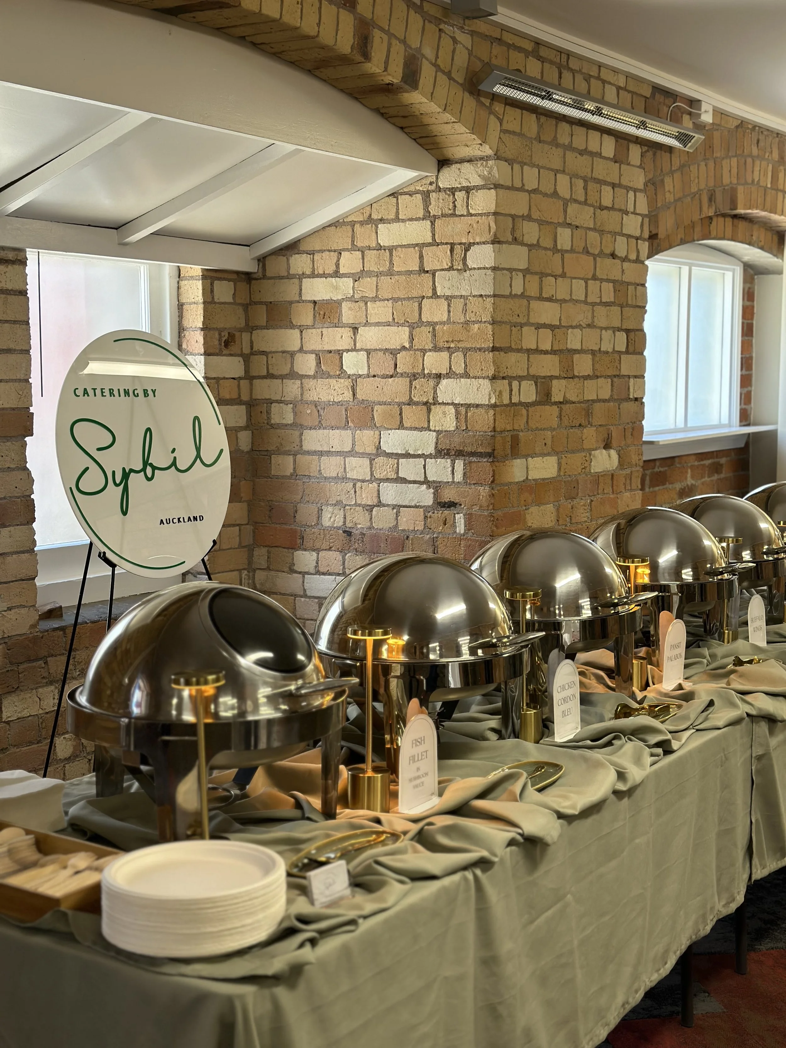 Buffet table with chafing dishes and plates, set up for catering in a room with brick walls and windows.