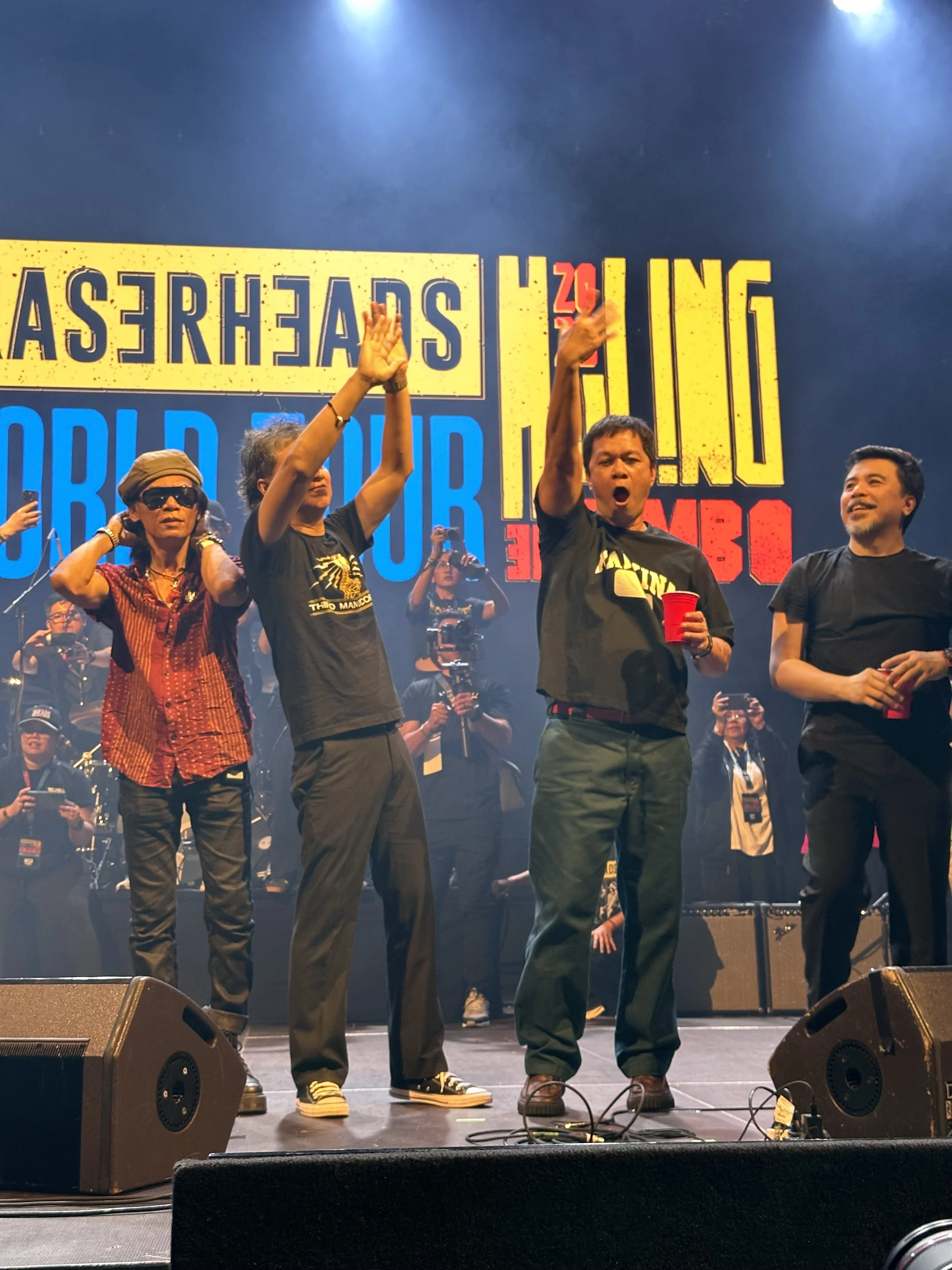 Group of musicians on stage performing at a concert with colorful background text. Some people are holding microphones and some are raising fists, dressed casually.