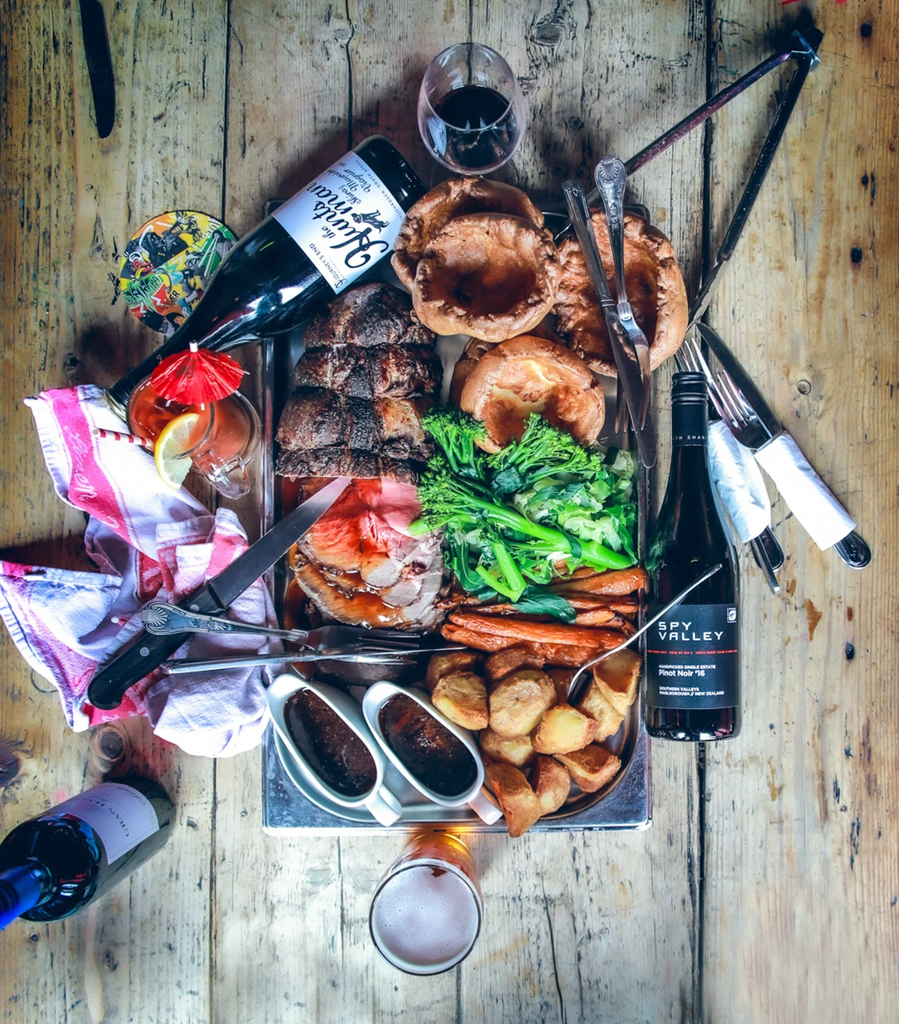 A spread of meat, vegetables, bread, and drinks on a wooden table for a meal.
