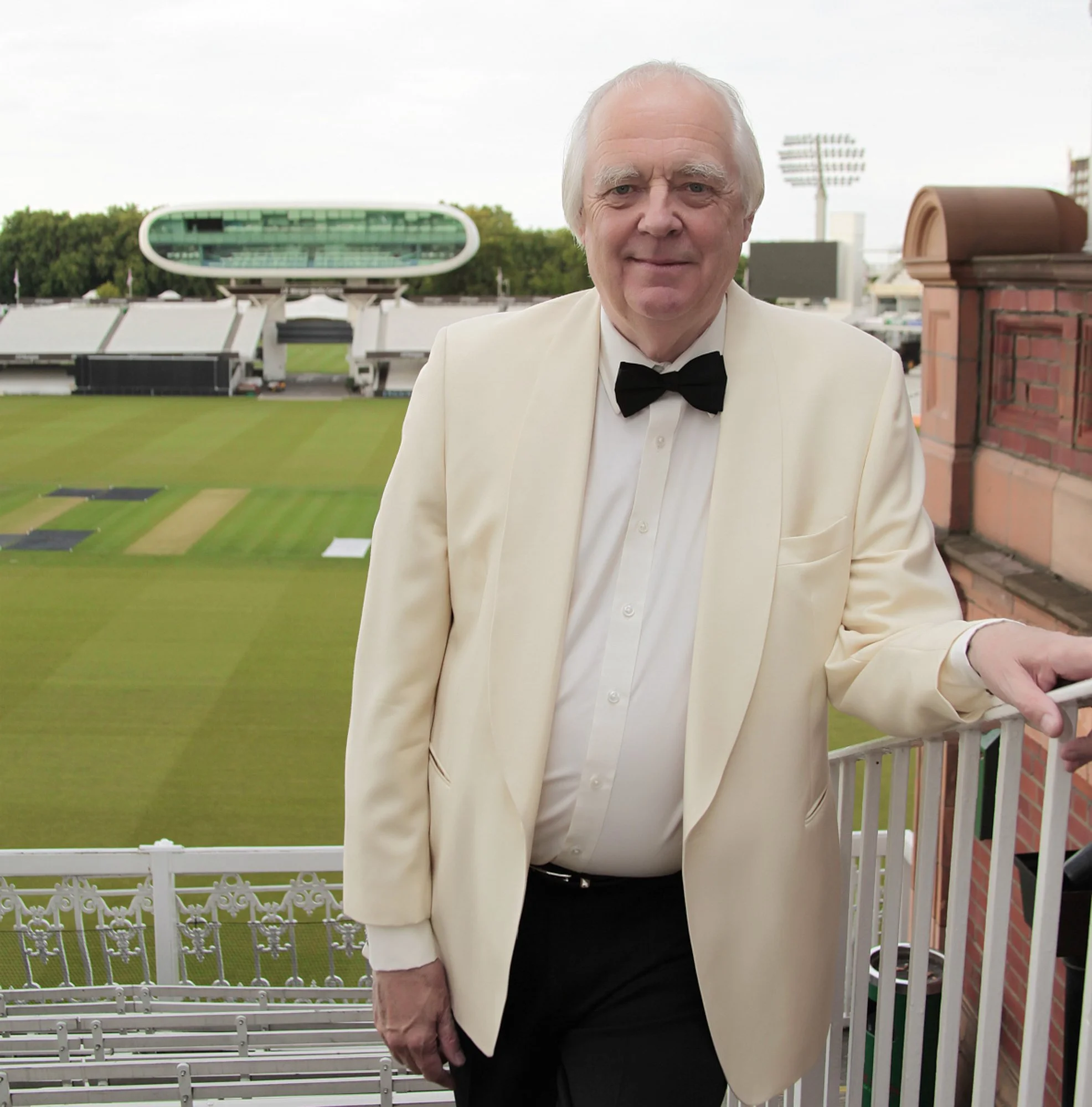 An elderly man dressed in a white tuxedo jacket, black bow tie, and white shirt standing on a balcony at a cricket stadium.