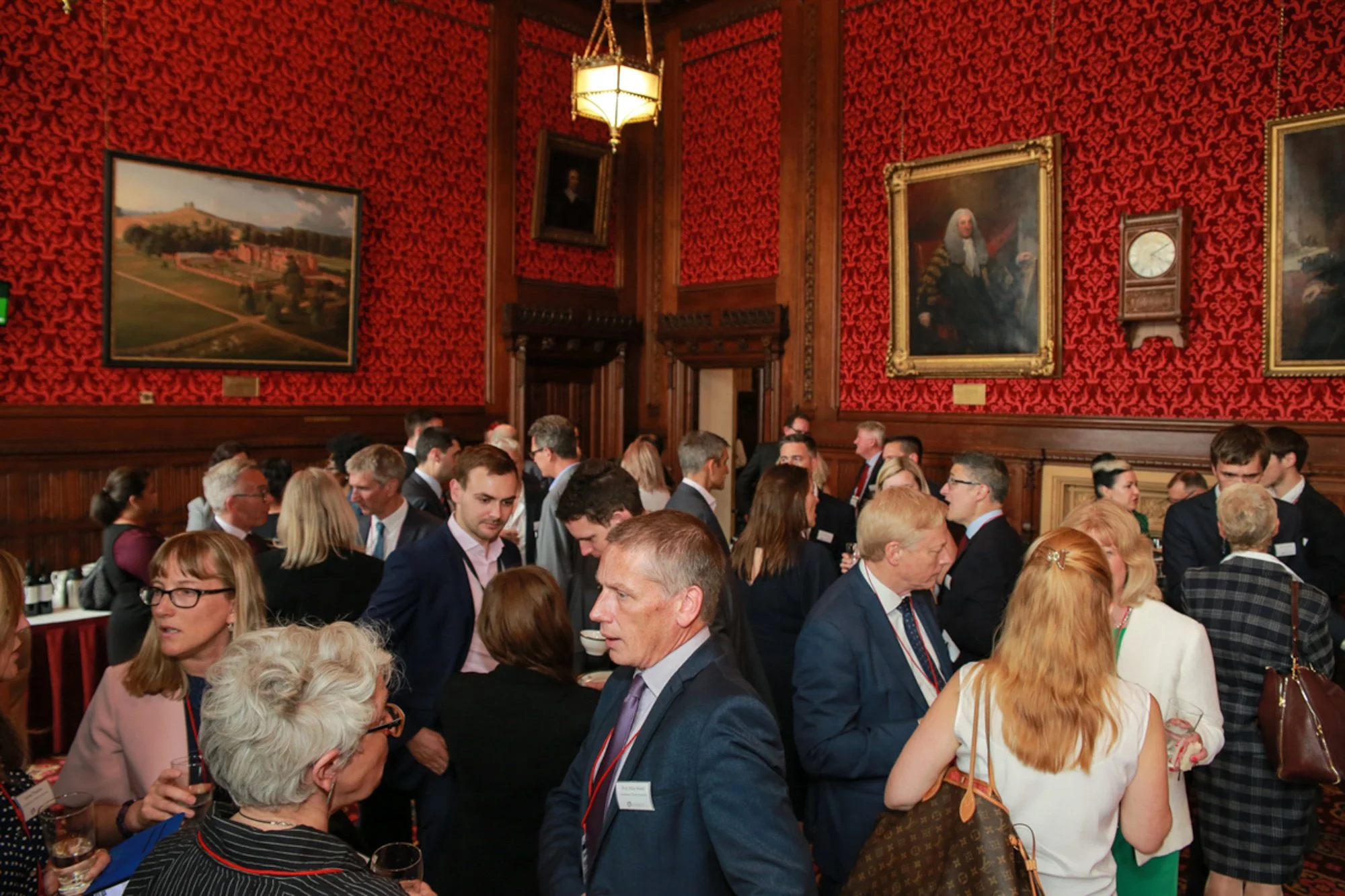 A crowded conference room with people socializing, with red ornate wallpaper, paintings, and a wooden-paneled interior.