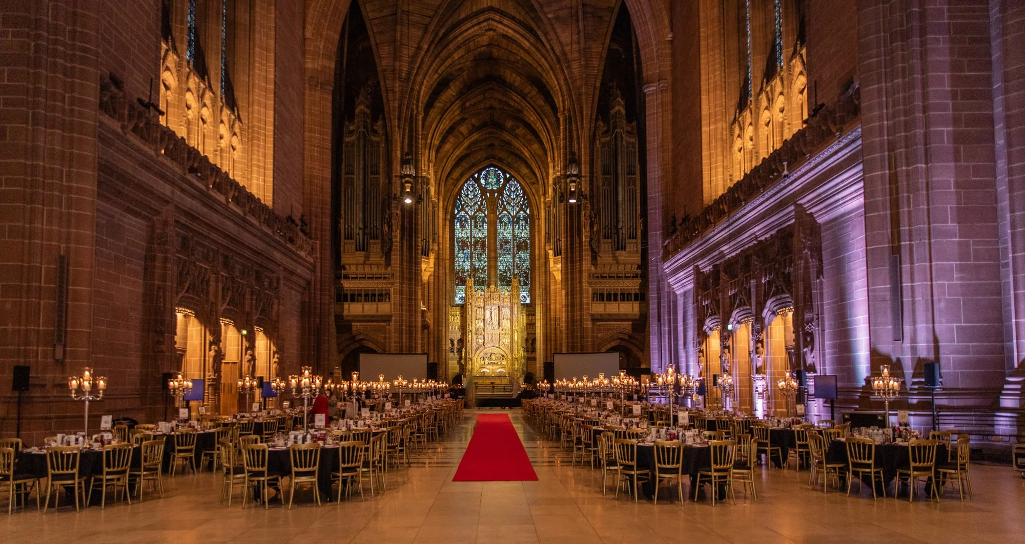 Elegant banquet hall inside a Gothic cathedral with high vaulted ceilings, stained glass windows, long dining tables with gold chairs, and candle centerpieces, illuminated in warm lighting.