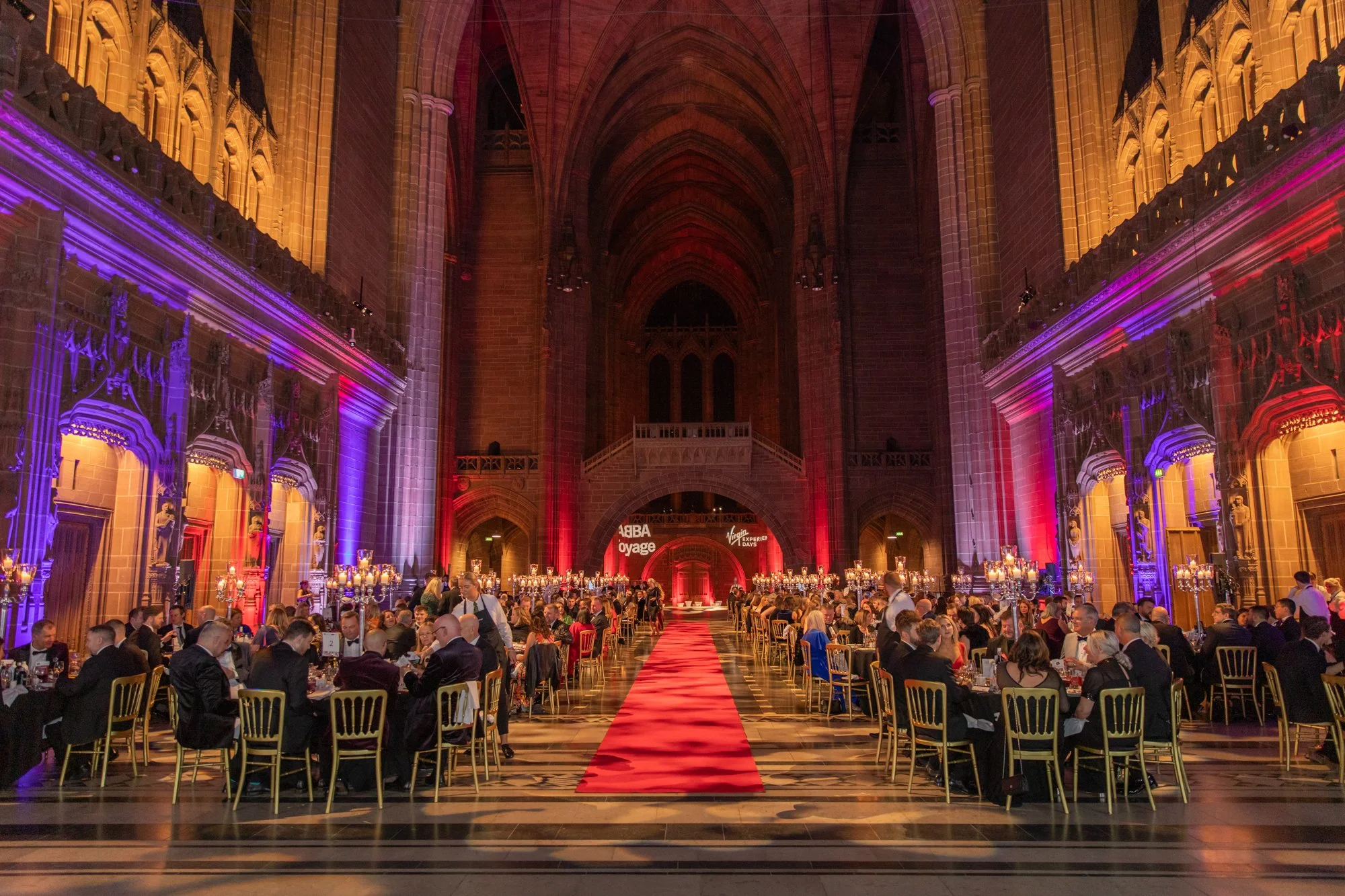 An elegant banquet hall with high arched ceilings, illuminated with purple and red lighting. Multiple round tables with black tablecloths and gold chairs are arranged around a red carpet runway in the center, leading towards a stage area with a backd