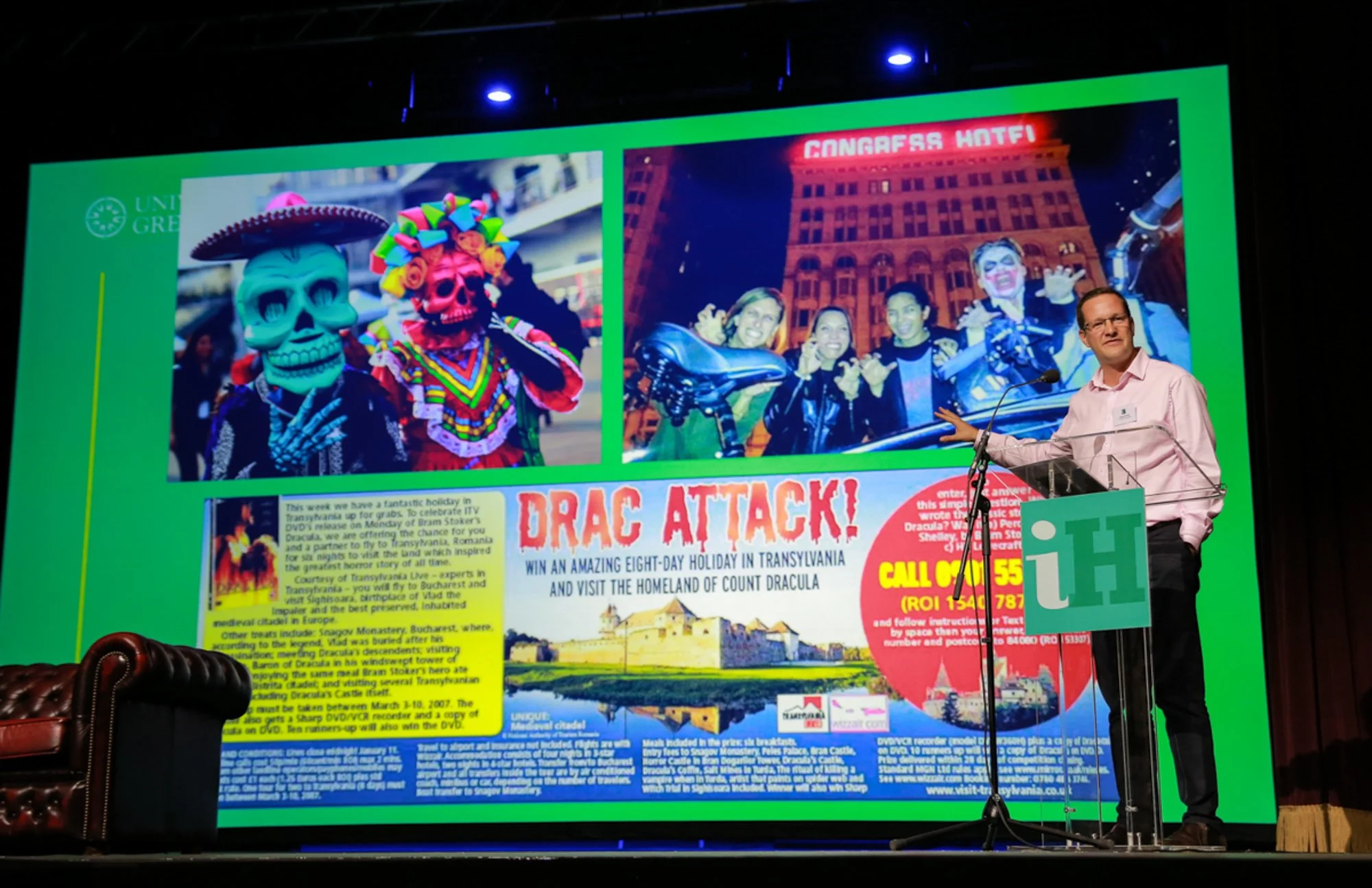 Man in pink shirt giving a presentation on a stage with a large colorful poster behind him promoting a Dracula-themed event in Transylvania, featuring images of people in costumes and promotional text.