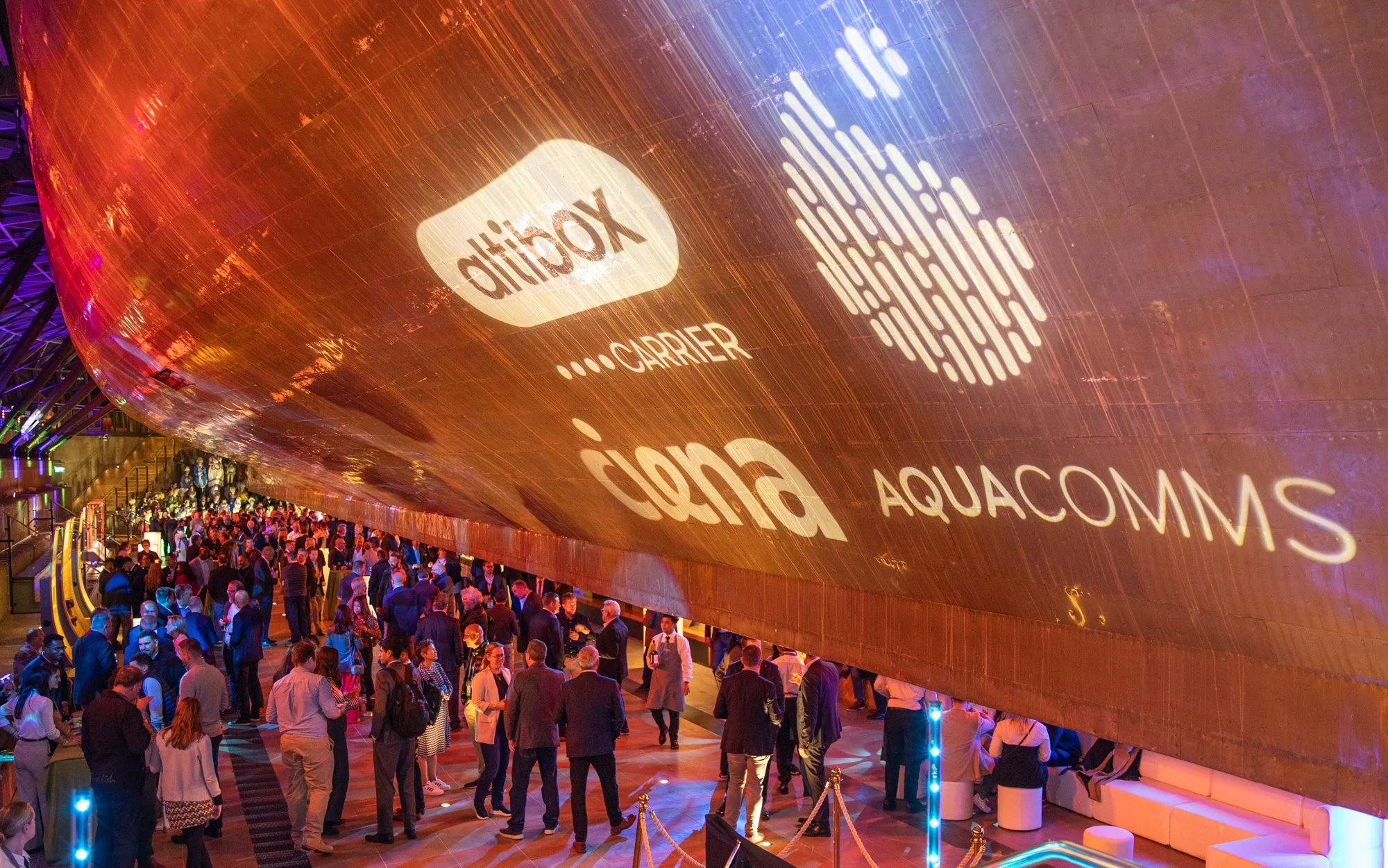 Large indoor event with many people mingling beneath a floating structure displaying logos of Vodafone, Cisco, and Aquacomms projected on its underside.