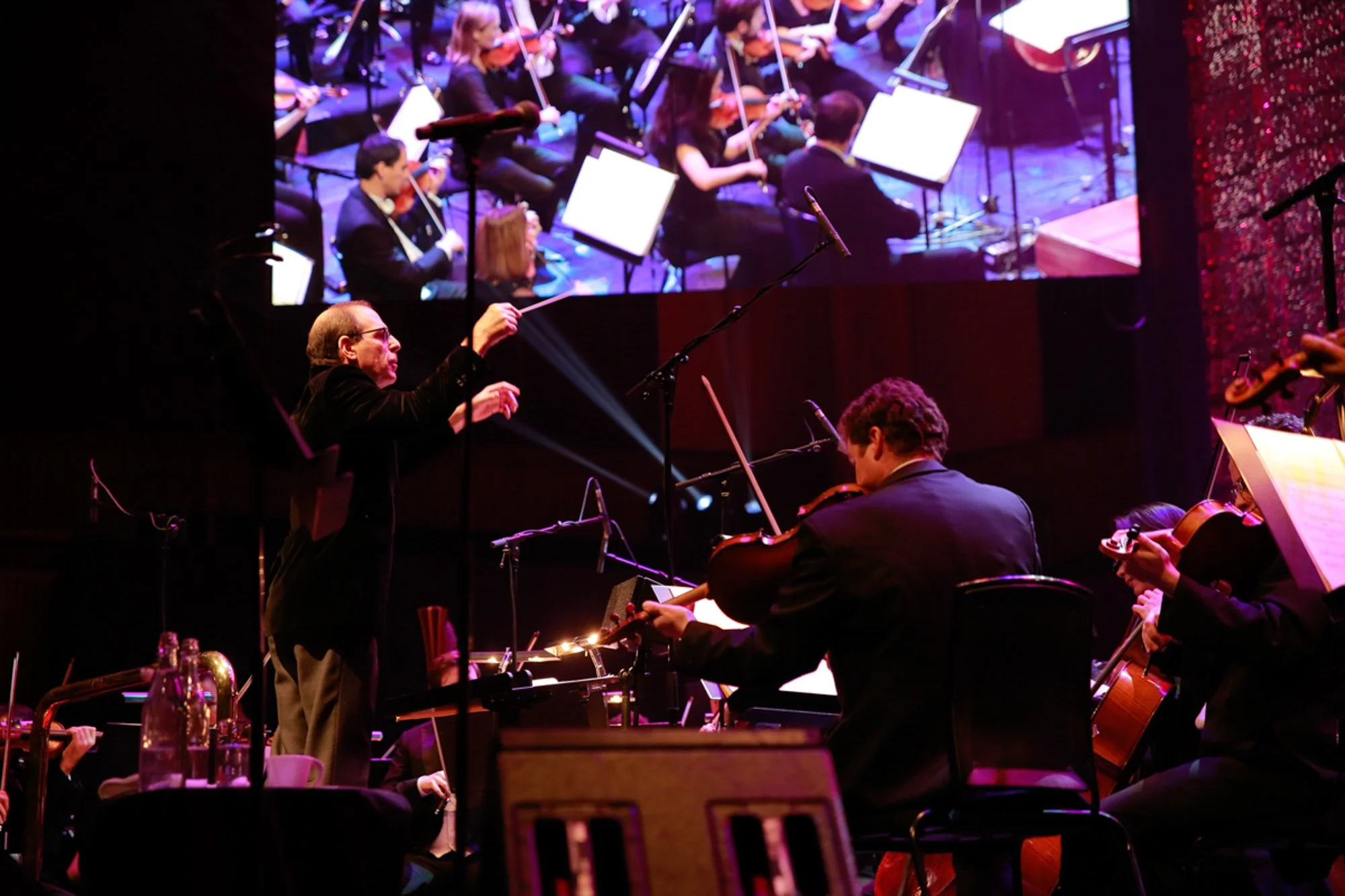 An orchestra performing on stage, with a conductor leading and musicians playing violins and other string instruments. A large screen above shows a close-up of the ensemble.