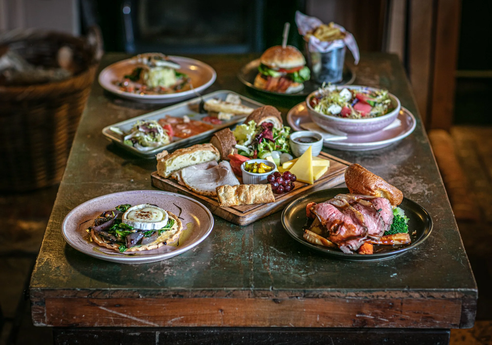 An assortment of dishes including salads, a burger, roast beef, a charcuterie platter with cheeses and grapes, and a dessert on a rustic wooden table in a cozy restaurant setting.