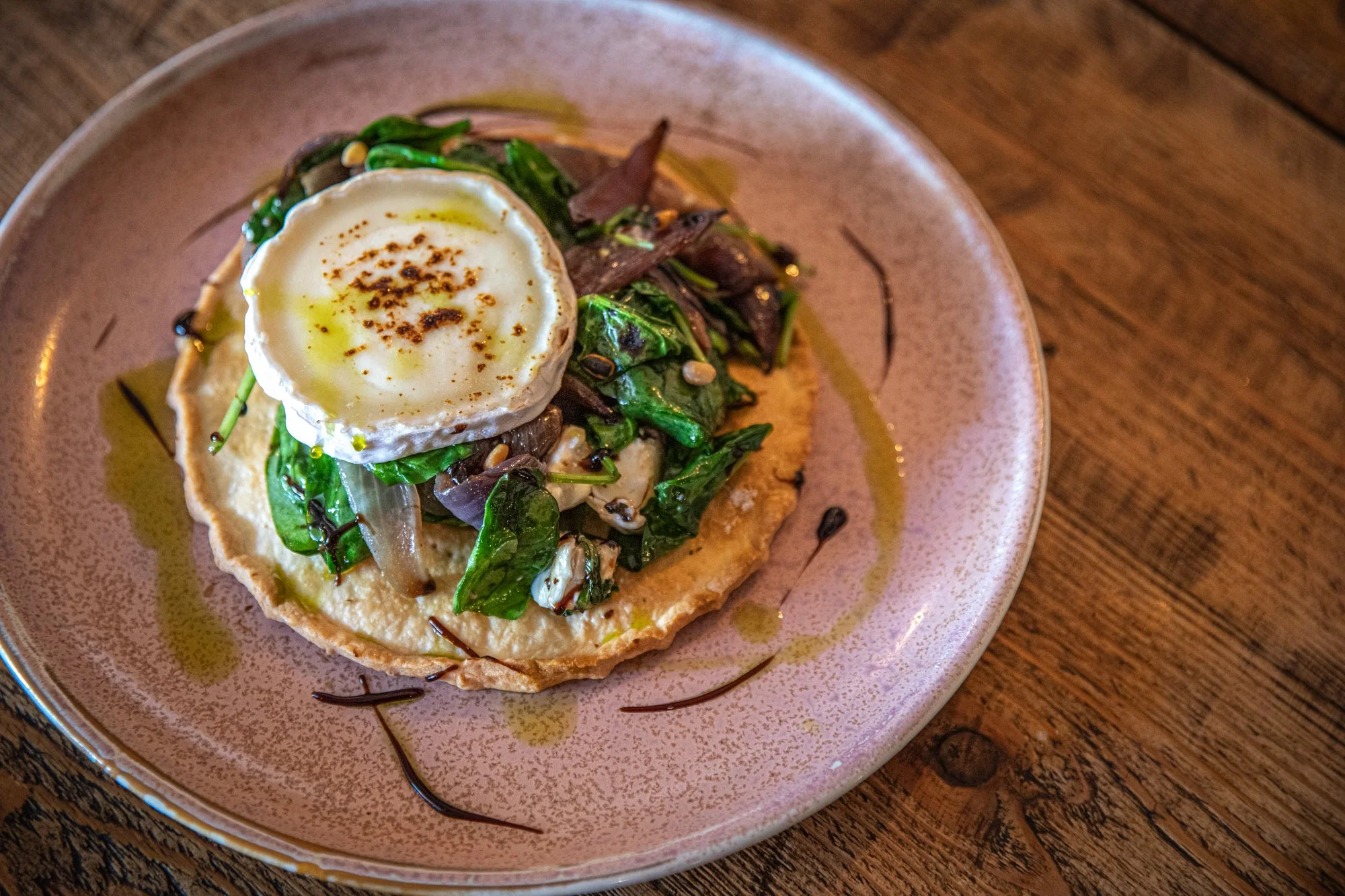 A dish with a baked egg on a bed of sautéed greens on top of mashed potatoes, lying on a rice paper crust, all drizzled with olive oil, served on a pink ceramic plate on a wooden table.