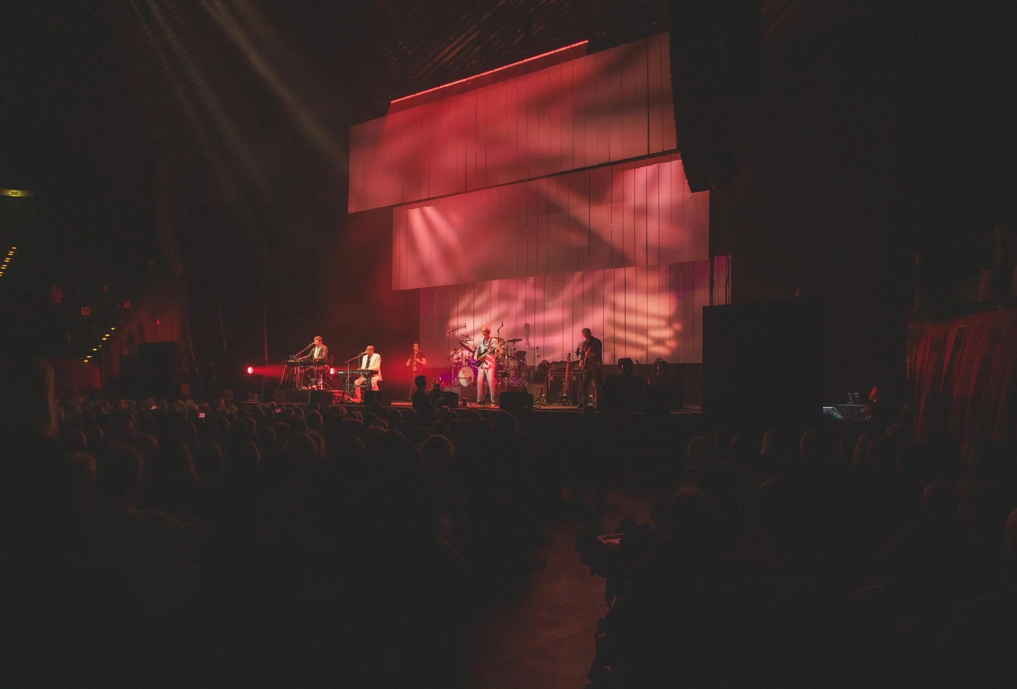A live band performing on stage with pink lighting and abstract visual background, audience seated in front.