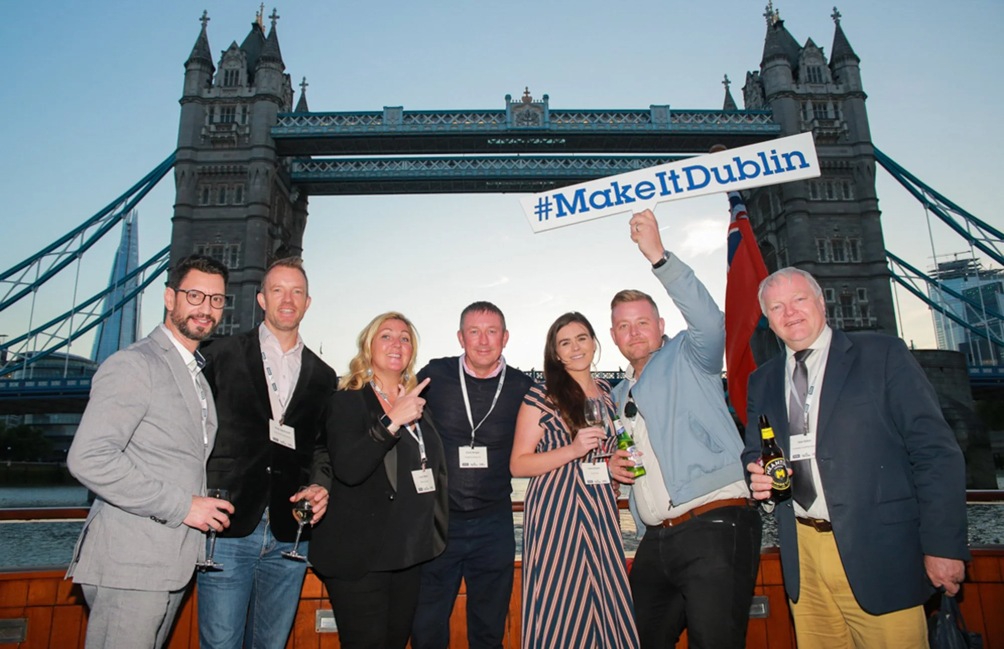 Group of seven people standing in front of Tower Bridge in London, holding drinks and a sign that reads #MakeItDublin.