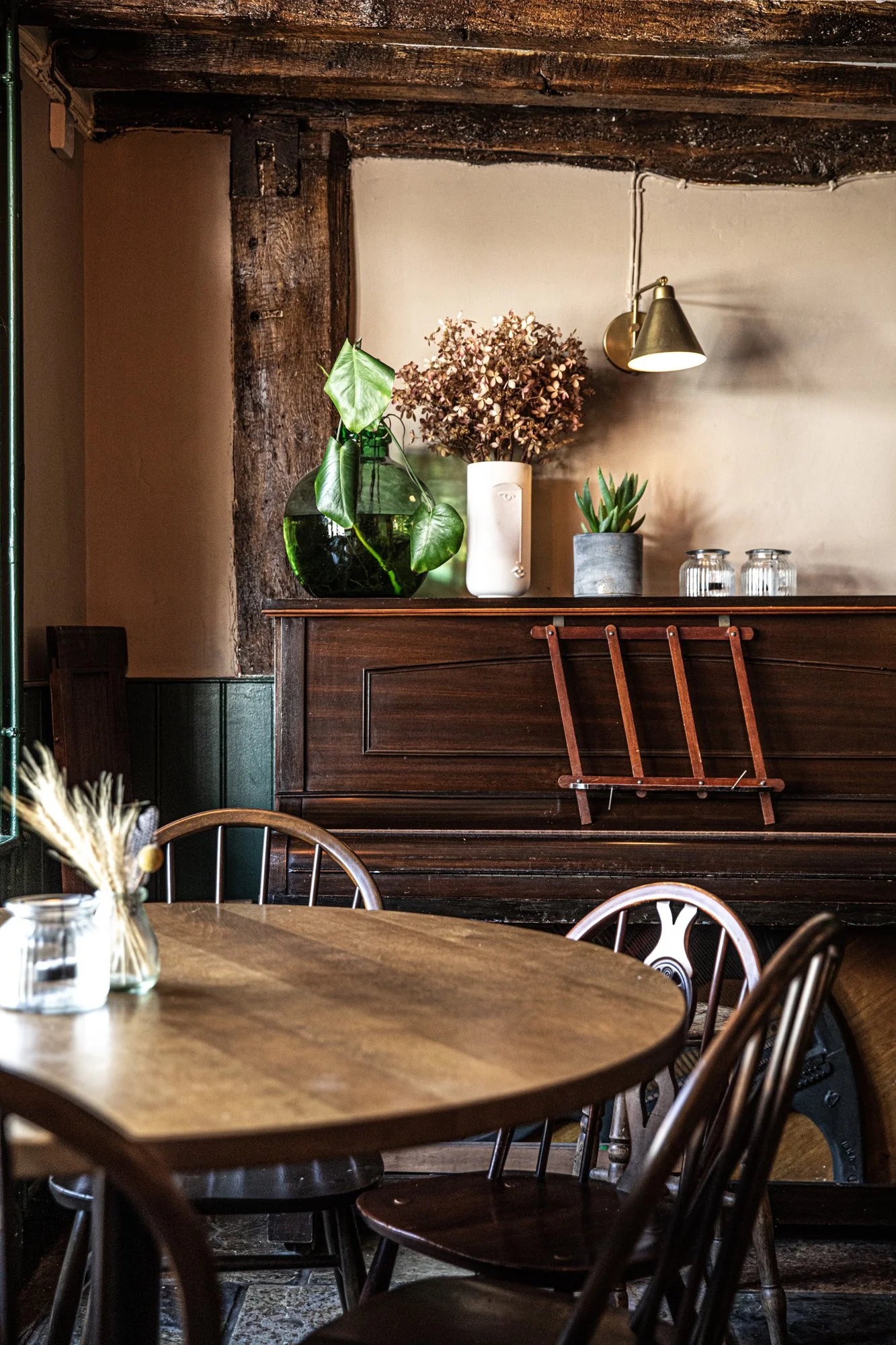 Interior of a cozy cafe with a wooden table and chairs, a piano decorated with potted plants and a wall-mounted light.