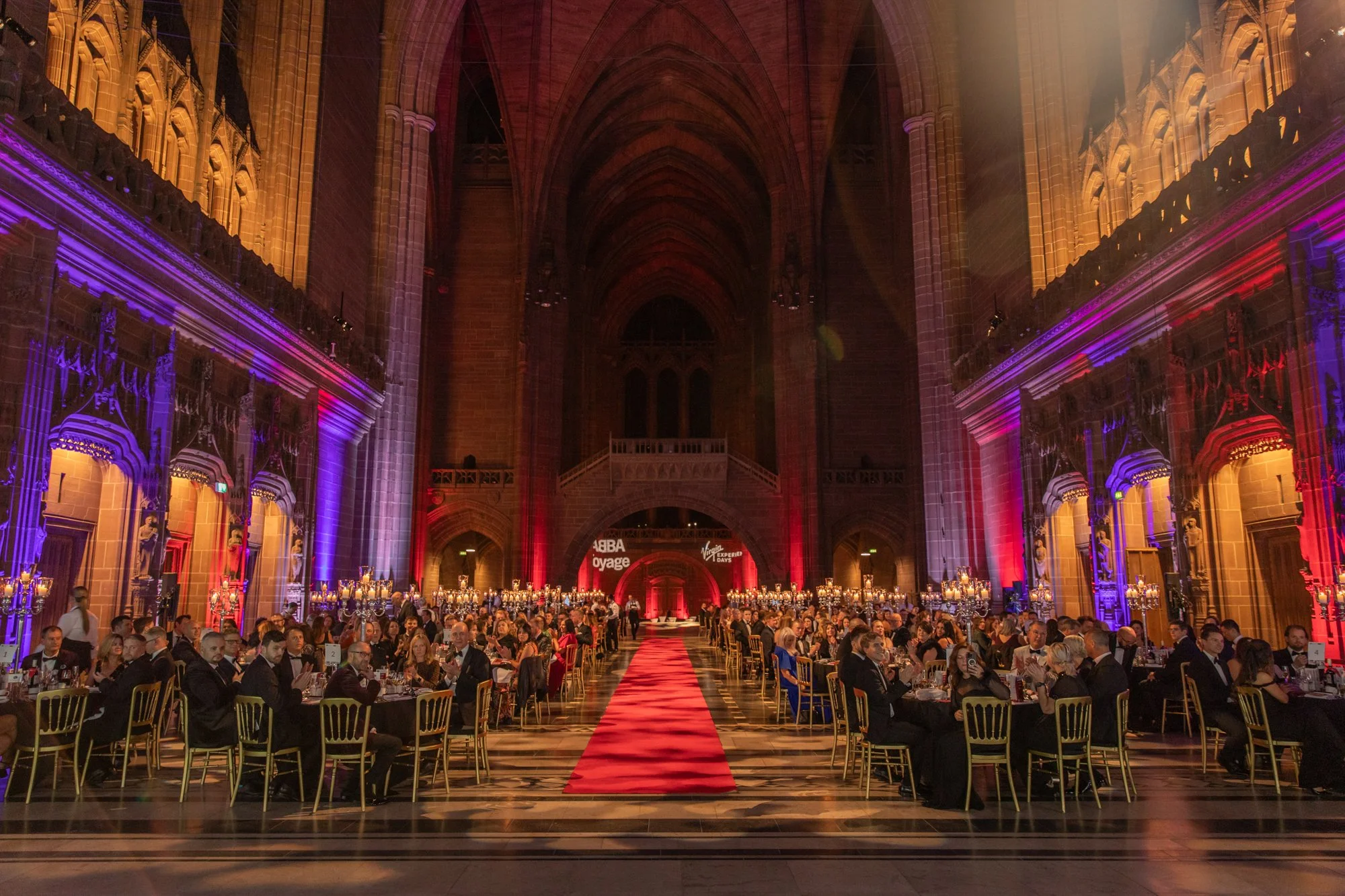 An elegant banquet in a grand hall with high arched ceilings, illuminated with purple and red lighting. Guests are seated at round tables with candles and floral centerpieces, and a red carpet runs down the center of the hall.