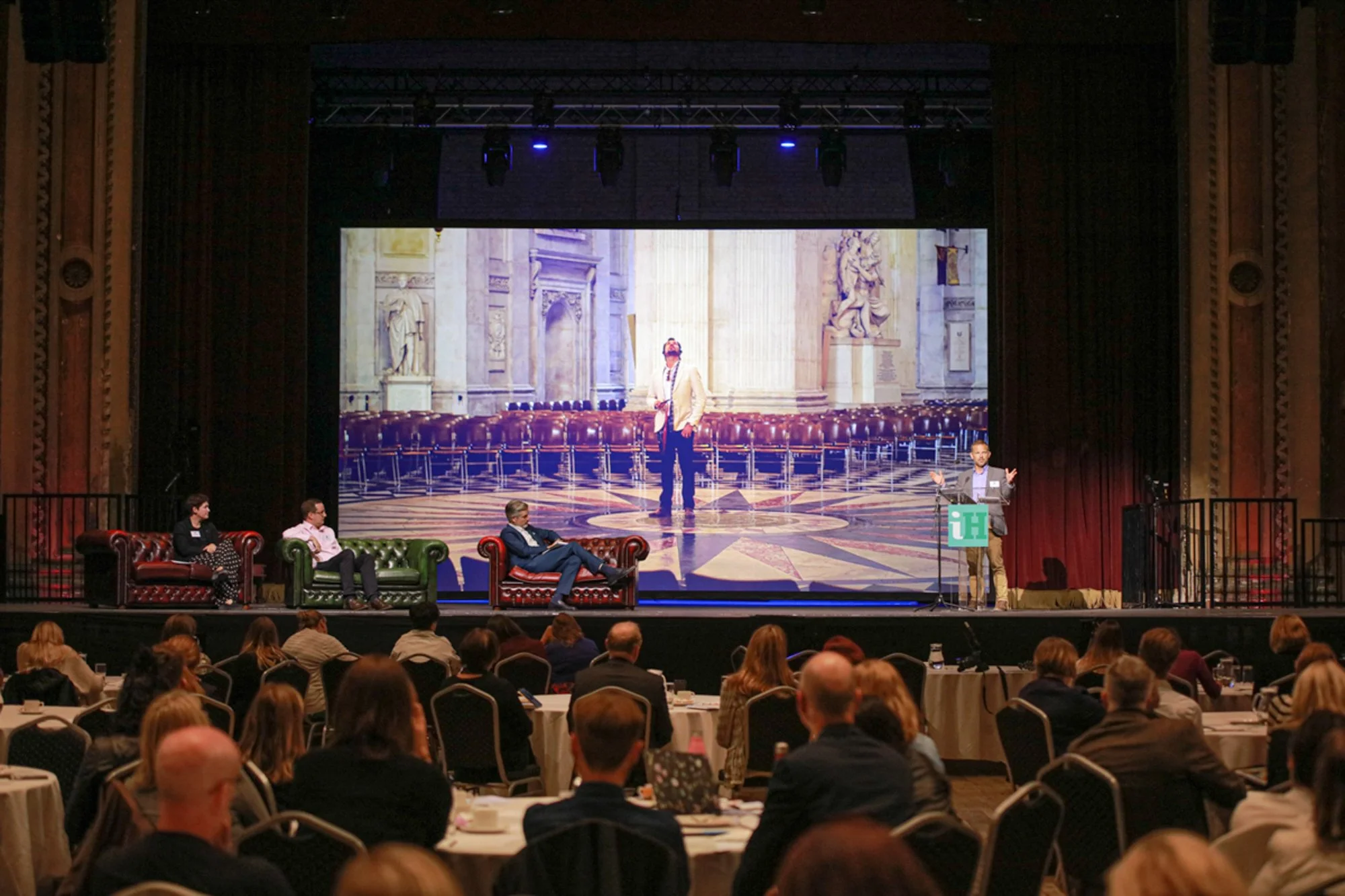 A conference or panel discussion taking place on a stage with three people seated on sofas and a speaker at a podium. Behind them, a large screen displays an image of a person standing inside a grand historical building with statues and ornate archit