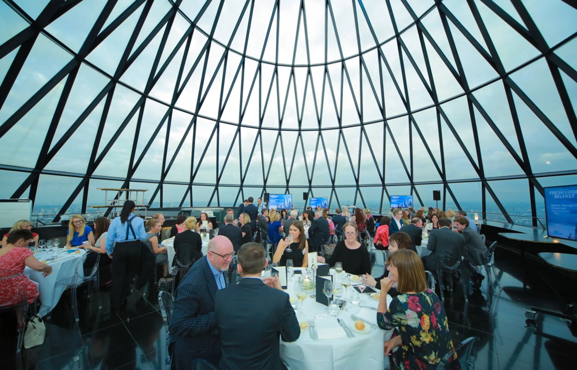 People attending a formal event or conference inside a glass-domed building with a view of the city skyline in the background. The interior has round tables set with glasses, plates, and silverware.