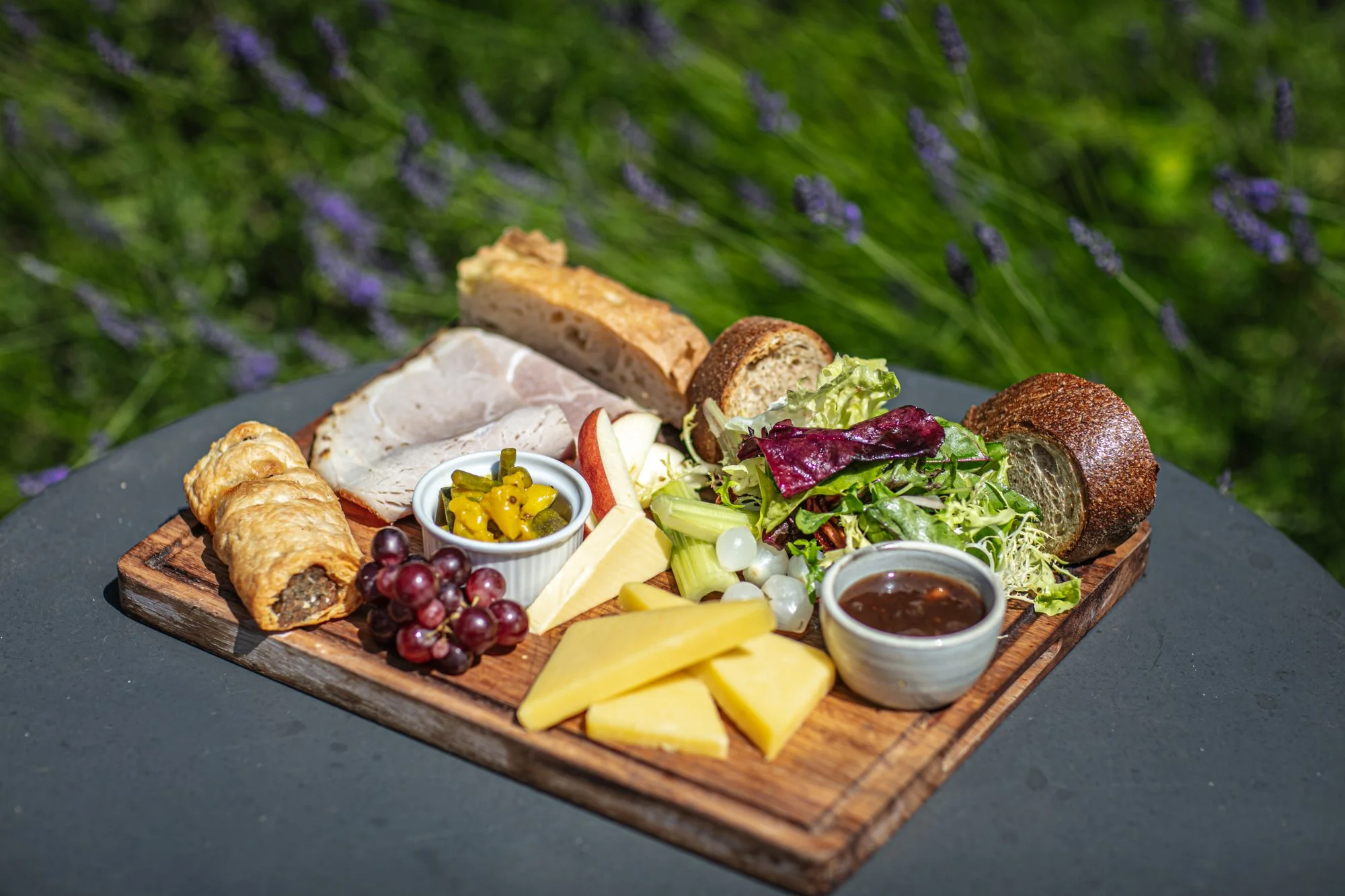 Assorted cheeses, fresh grapes, sliced apples, mixed greens salad, bread, crackers, fruit chutney, and jam on a wooden serving board outdoors with greenery in the background.