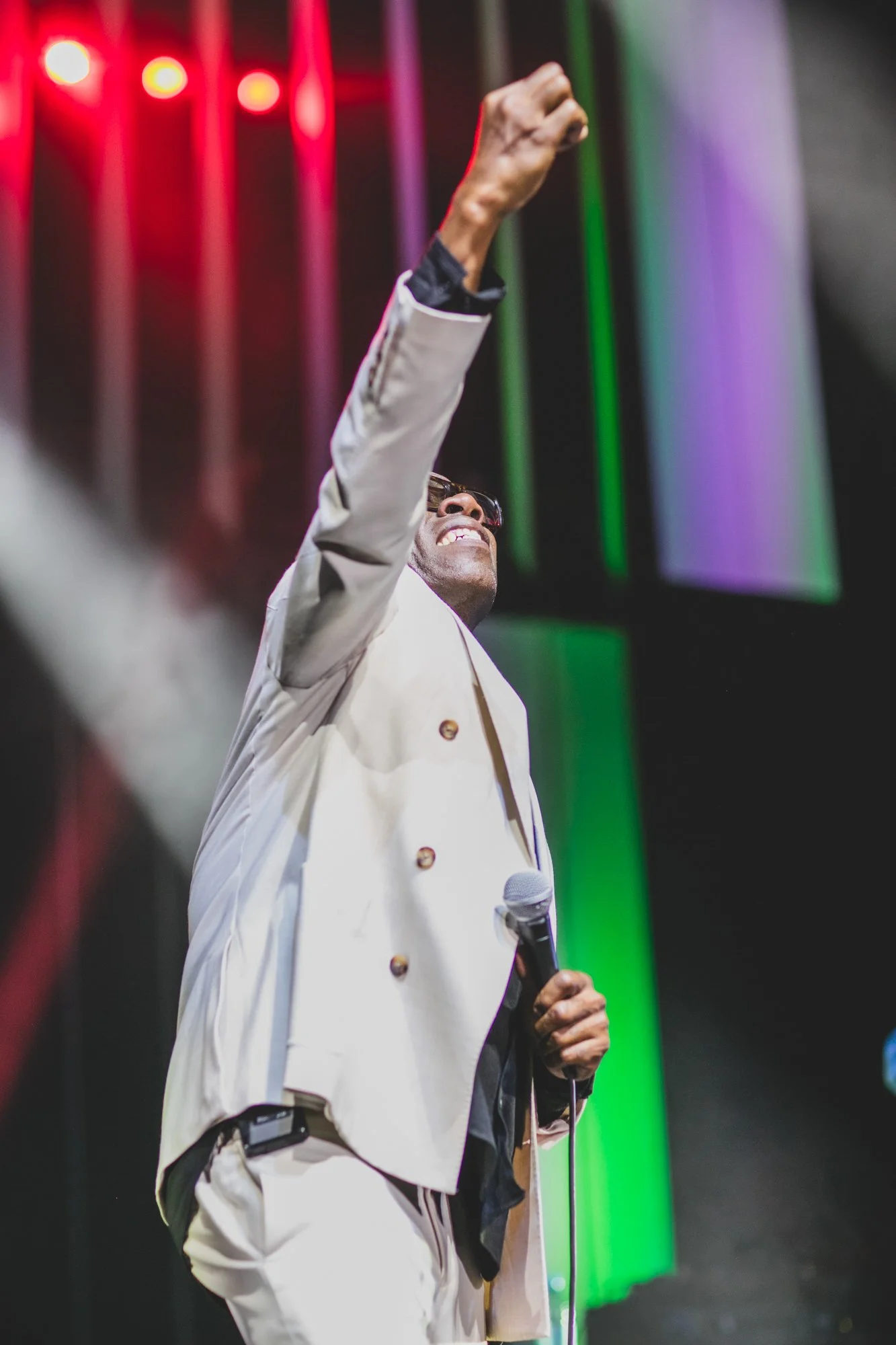 Person in a white suit with sunglasses holding a microphone and raising their fist on stage with colorful lights in the background.