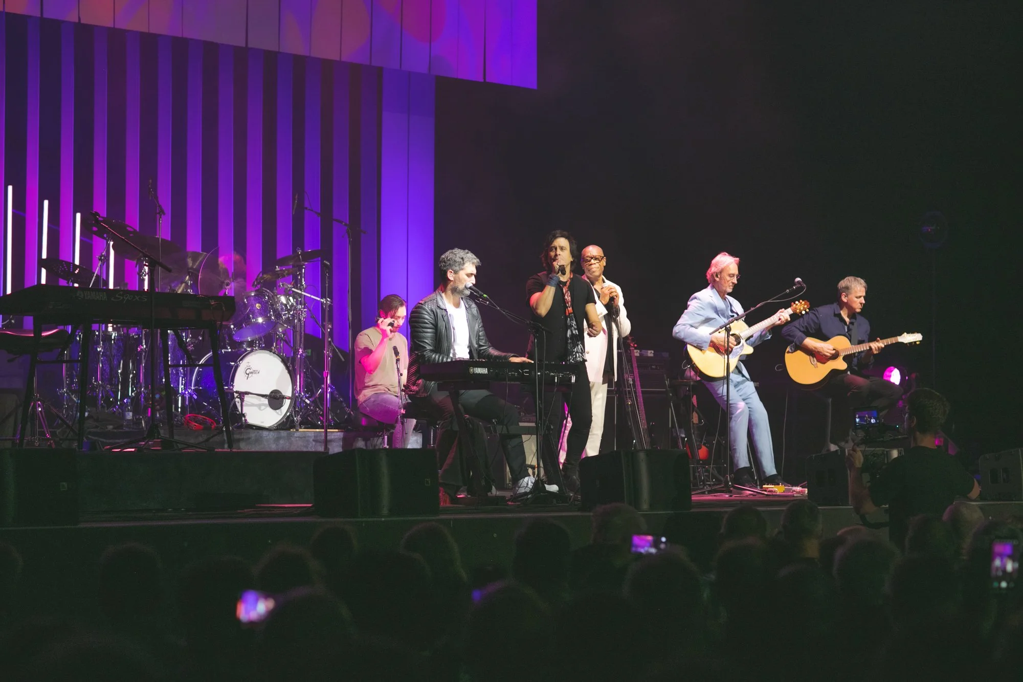 Musicians performing on stage during a concert with purple lighting, including a drummer, keyboardist, and guitarists, with a live audience watching.