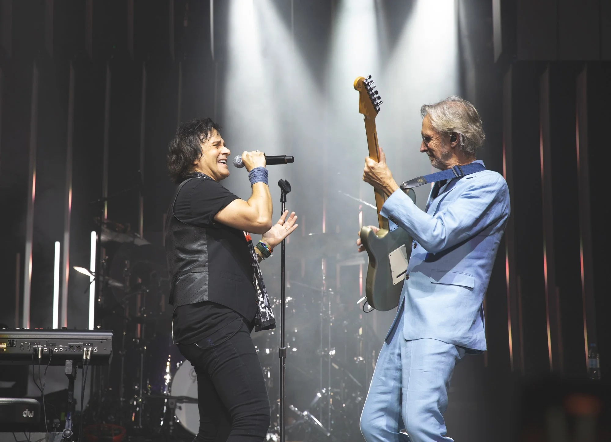 Two musicians perform on stage: a woman singing into a microphone and a man playing an electric guitar, with stage lights and musical equipment in the background.