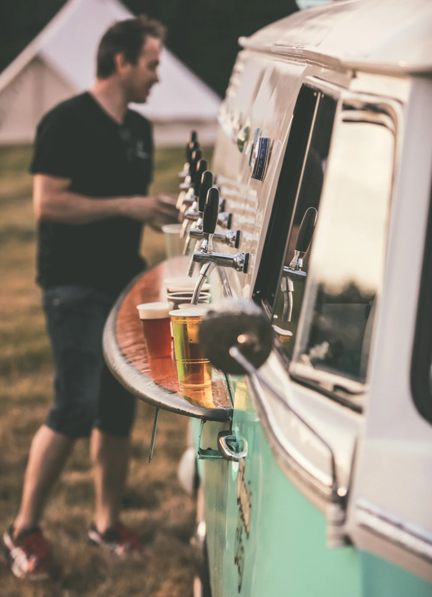 A man in a black shirt and shorts pours drinks from taps on a vintage-style drink truck with a wooden bar top, with cups of beer in front of him. The scene is outdoors during the evening or late afternoon.