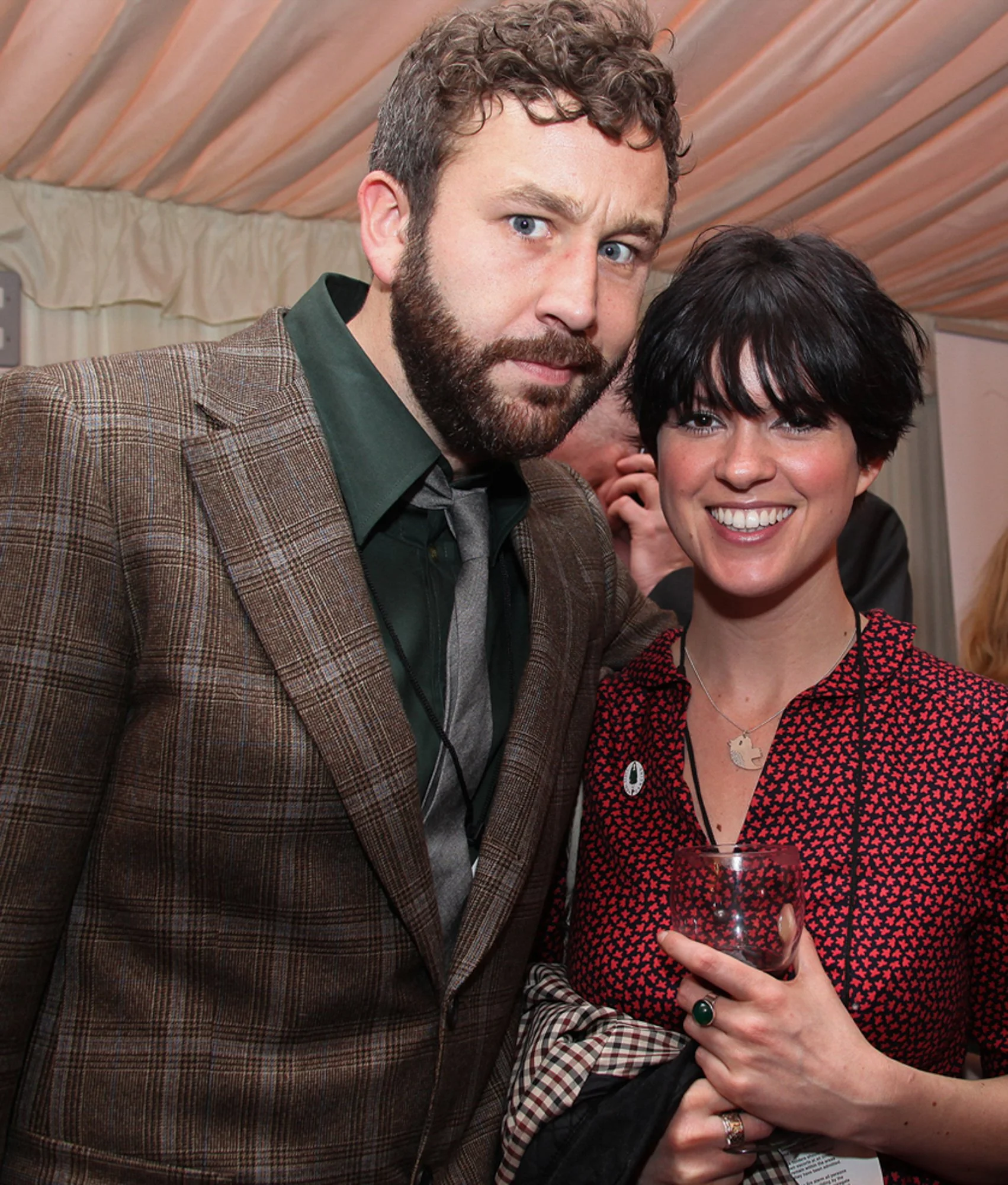 A man with curly hair, beard, wearing a checkered blazer and dark shirt standing next to a woman with short black hair, wearing a red and black patterned blouse, holding a glass of red wine, smiling at the camera.