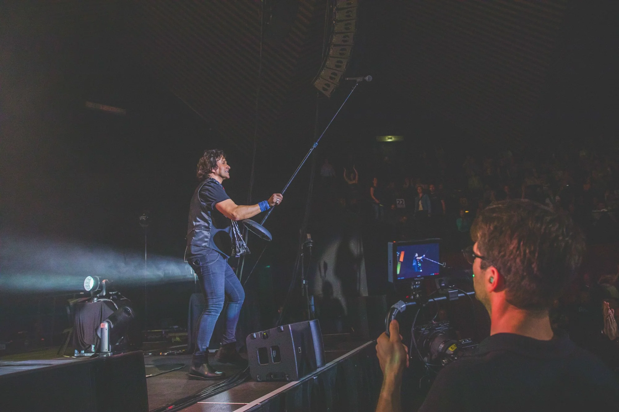 A man with long curly hair wearing a black shirt and jeans holding a microphone stand on stage, with a woman in the background raising her hands, and another man filming the performance with a video camera in a dark concert venue.