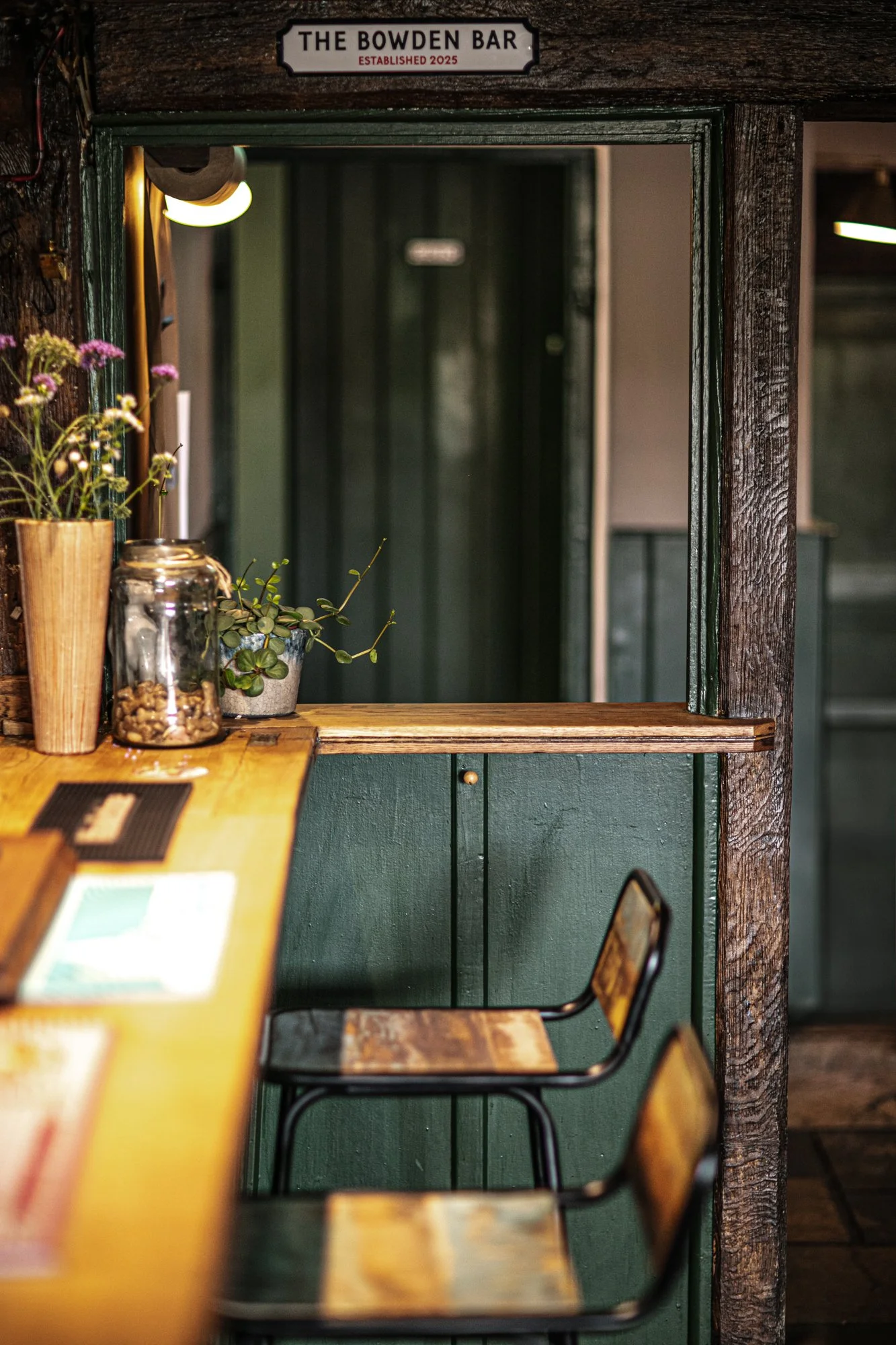 Interior of a bar with a wooden counter, potted plants, and chairs. A sign above the window reads 'The Bowden Bar, Established 2025'.