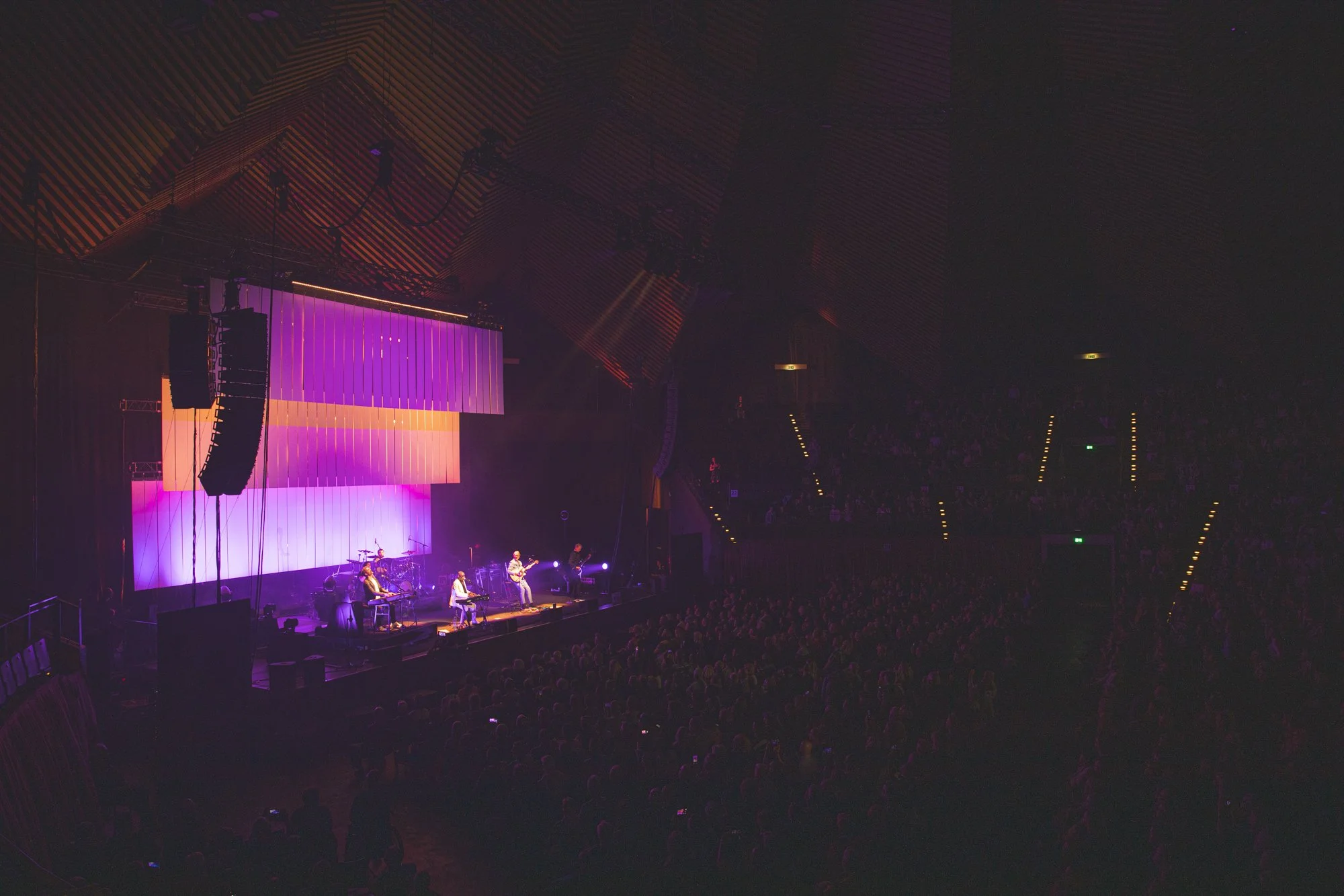 A live concert on a stage with purple and orange lighting, featuring a band playing instruments and a large audience in a darkened auditorium.