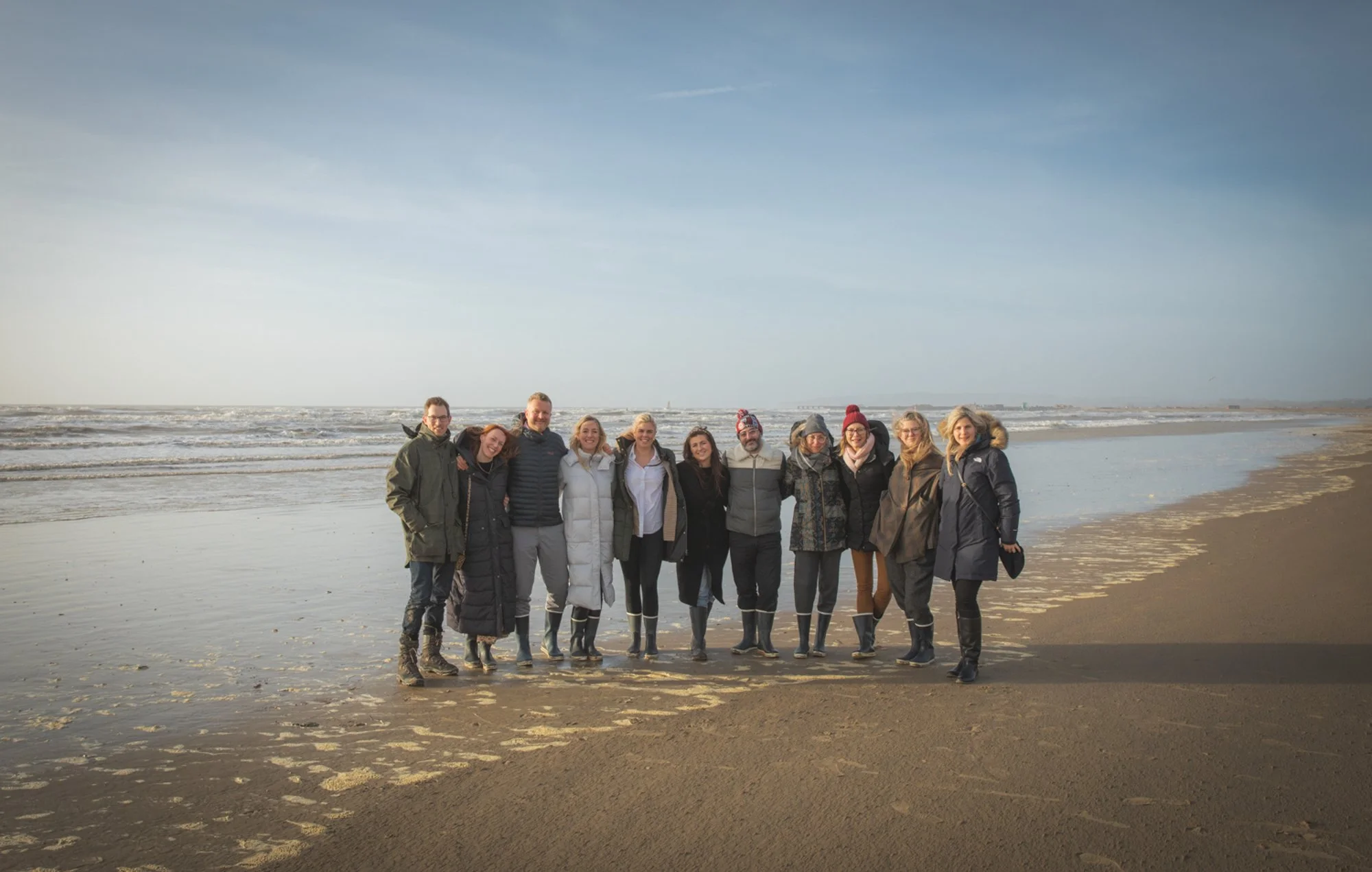 Group of eleven people standing on the beach near the ocean, dressed warmly with jackets and boots, smiling and posing for the photo.