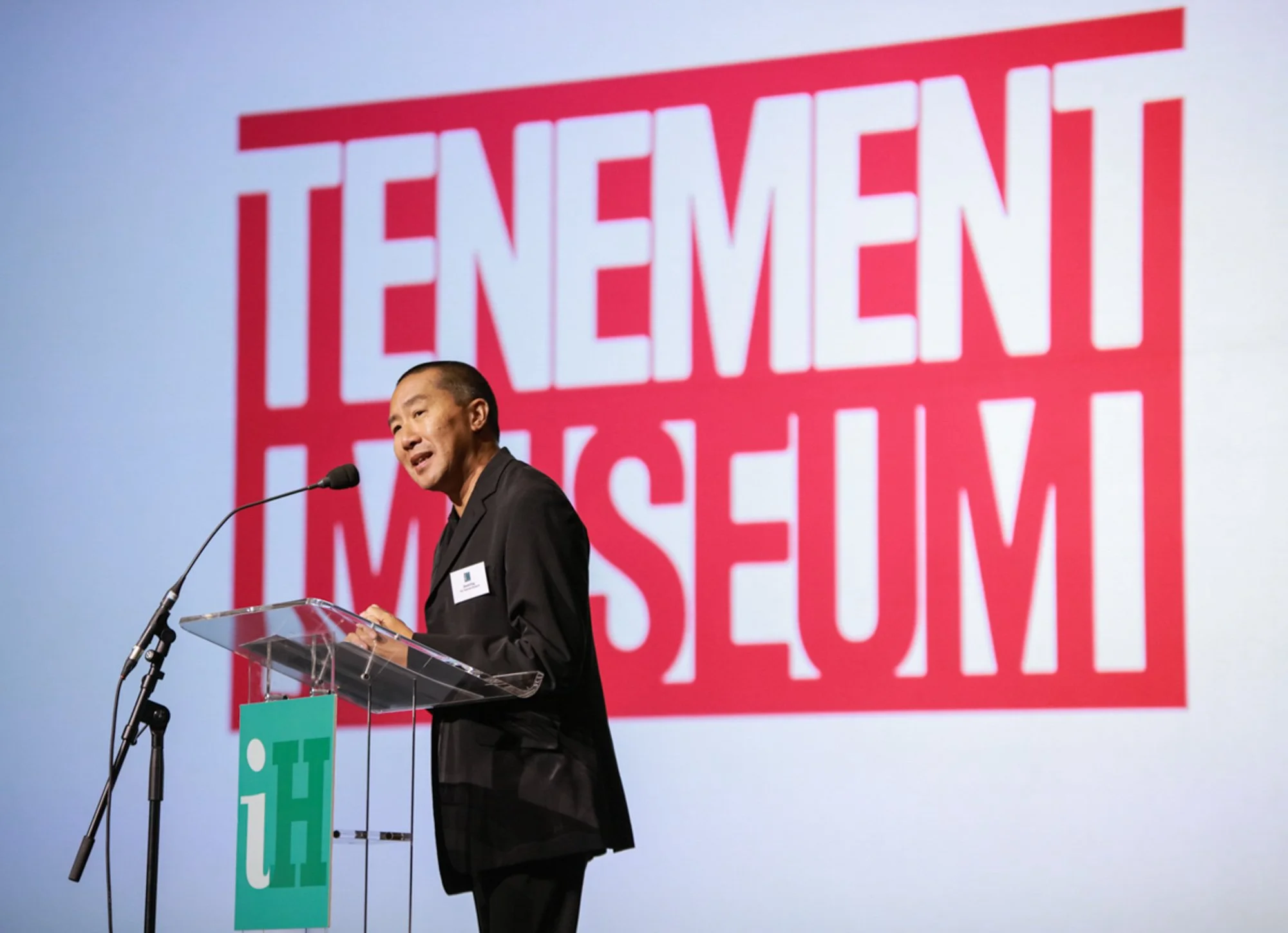 Speaker at a podium with a large screen behind him displaying the words 'Teniem Rent Meseum' in red text.