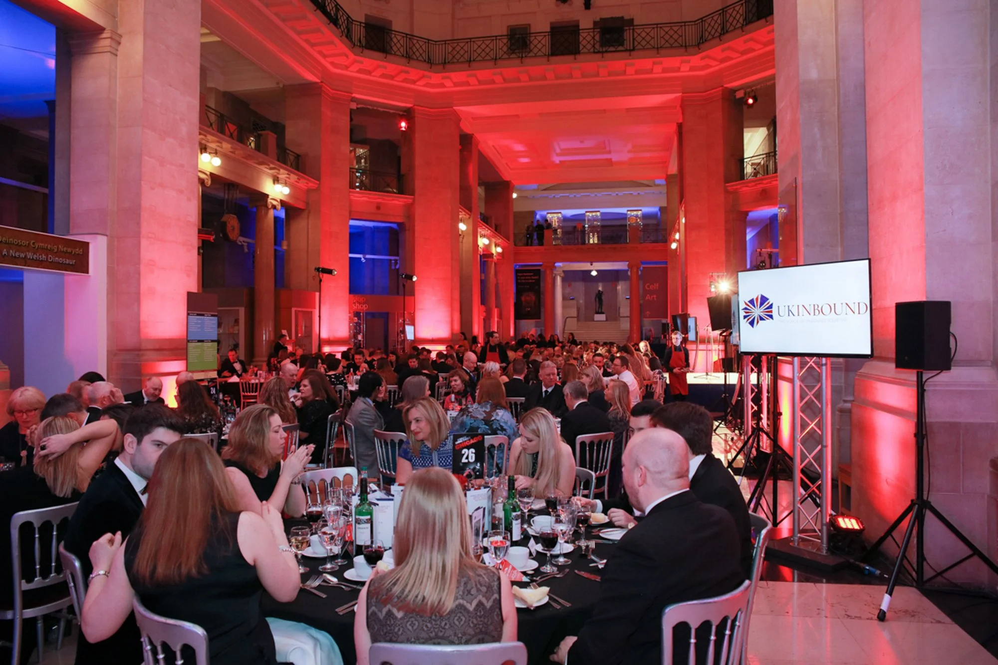 A large formal gathering of people seated at round tables in a grand hall with high ceilings, illuminated with red and blue lighting, and a large screen displaying the logo 'UKINBOUND'.
