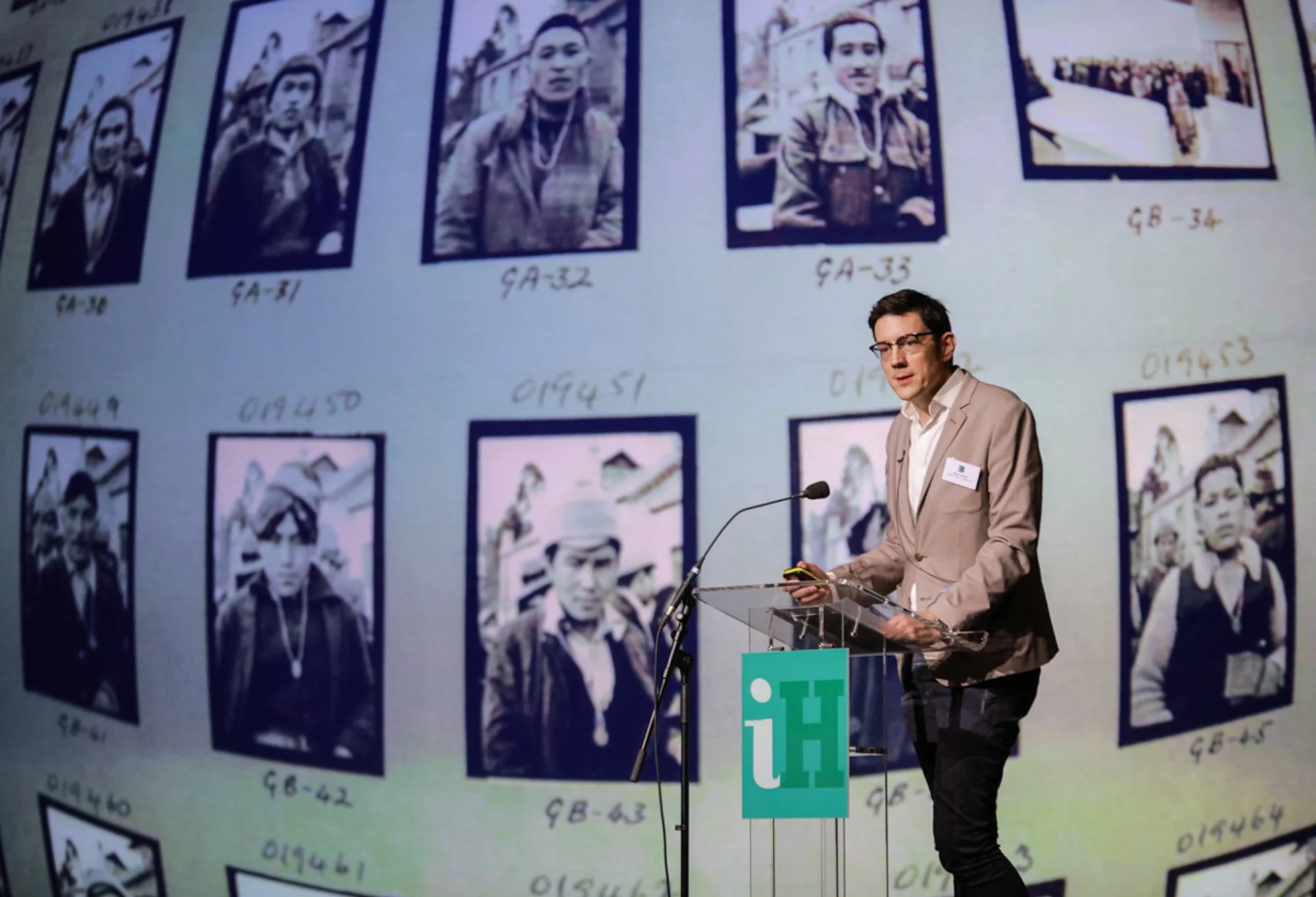 A man in a beige blazer and glasses stands at a podium with a microphone, giving a presentation. Behind him, a large screen displays black-and-white photographs of people, each labeled with a code.