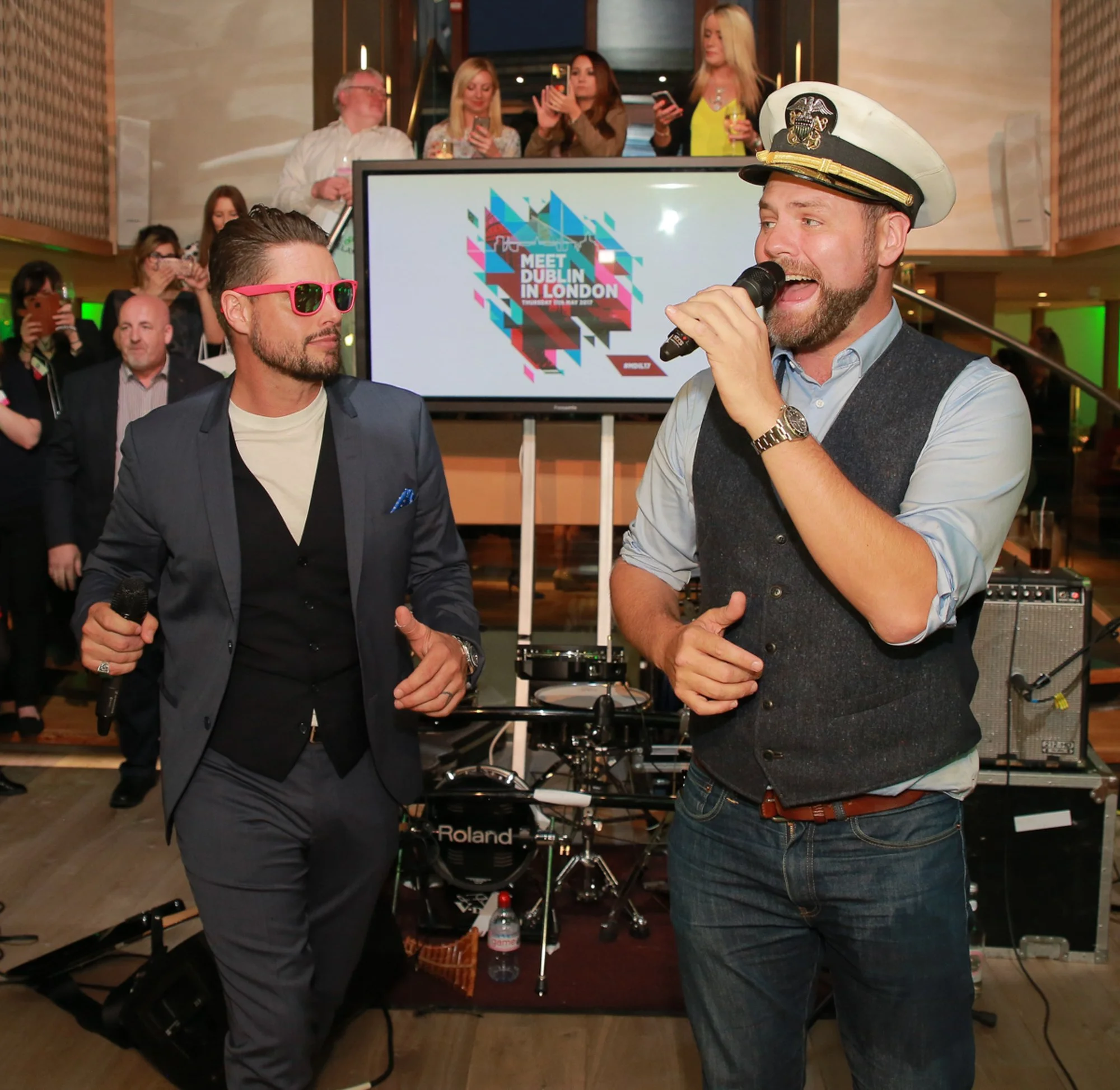 Two men speaking into microphones at an indoor event with a crowd, a DJ setup, and a digital screen in the background displaying "Meet Dublin in London".