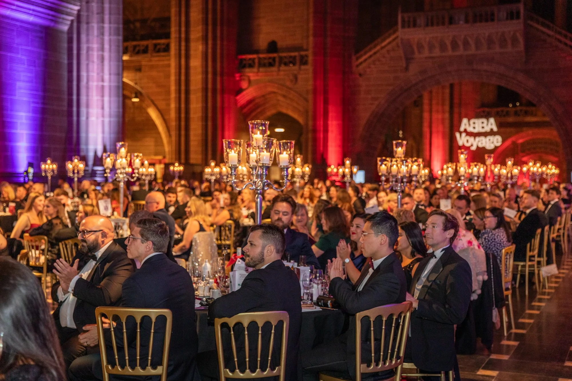 People dressed in formal attire sitting at banquet tables during a formal event inside a grand hall decorated with chandeliers and lighting, with a projection saying 'ABBA Voyage' in the background.