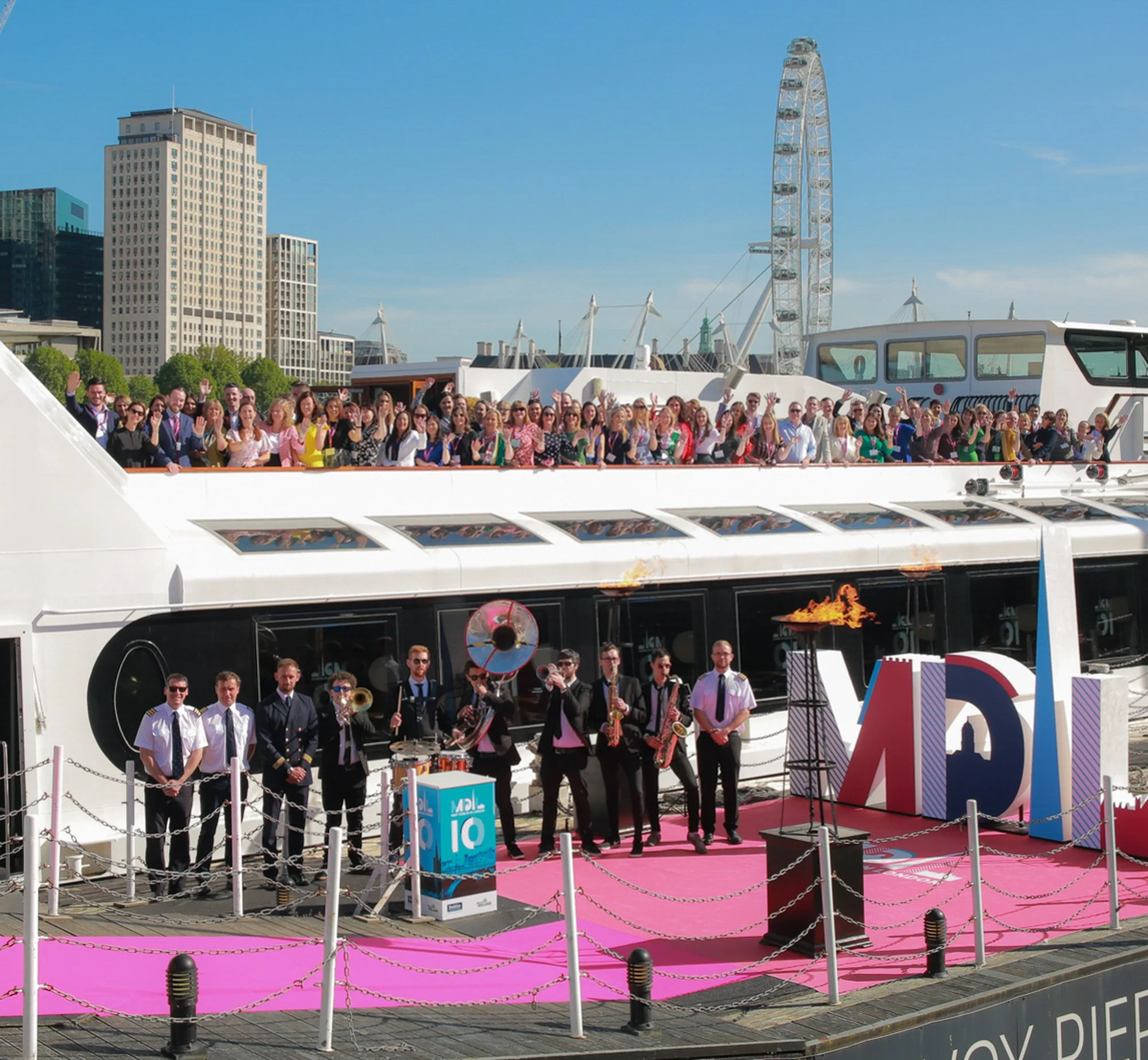 A large group of people on a boat and dock celebrating during daylight, with a band playing music and the London Eye visible in the background.