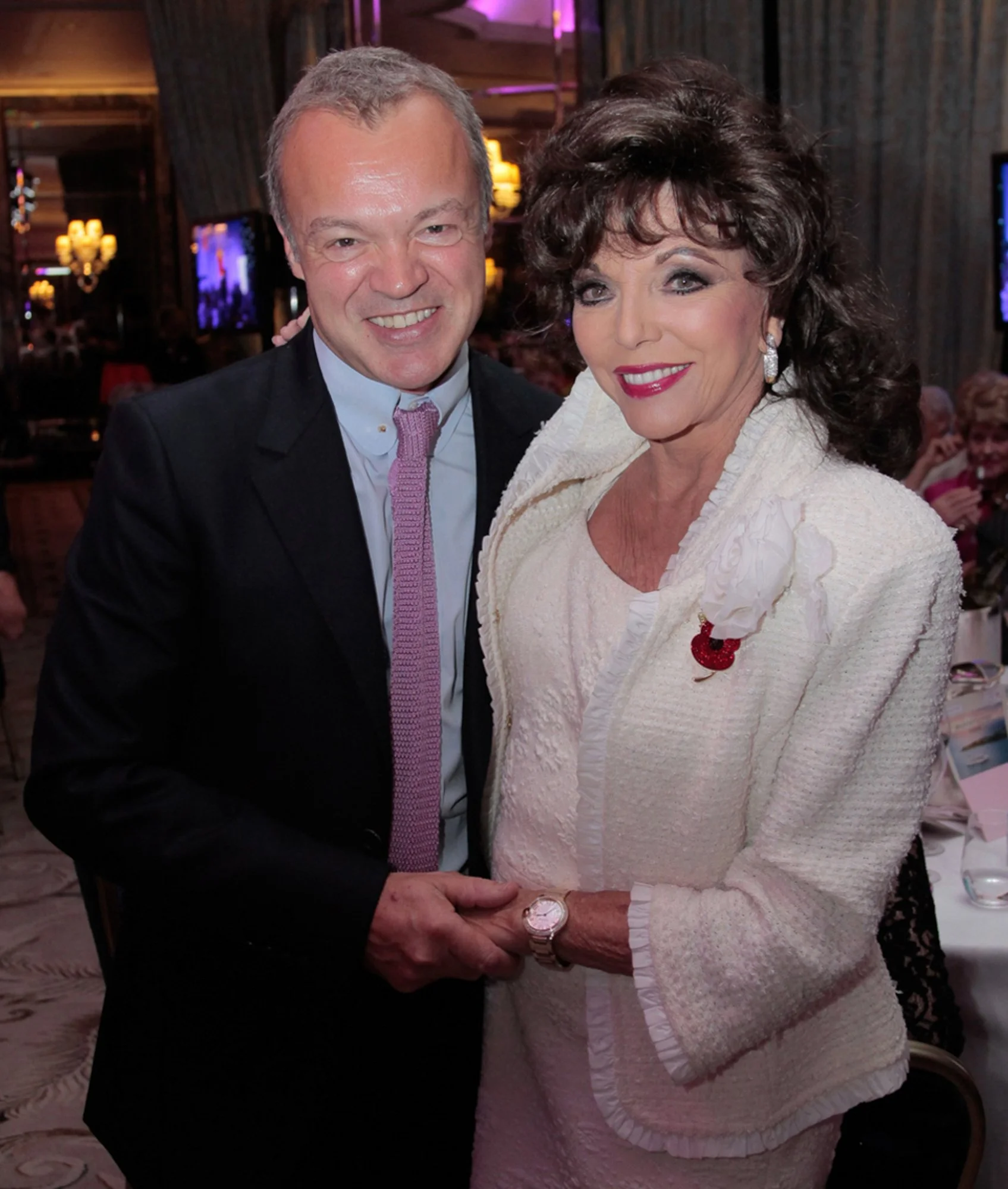 A man and a woman are smiling and holding hands at an indoor event. The man is wearing a black suit with a white shirt and a pink tie, and the woman is wearing a cream-colored textured suit with a flower brooch, earrings, and a watch. The background