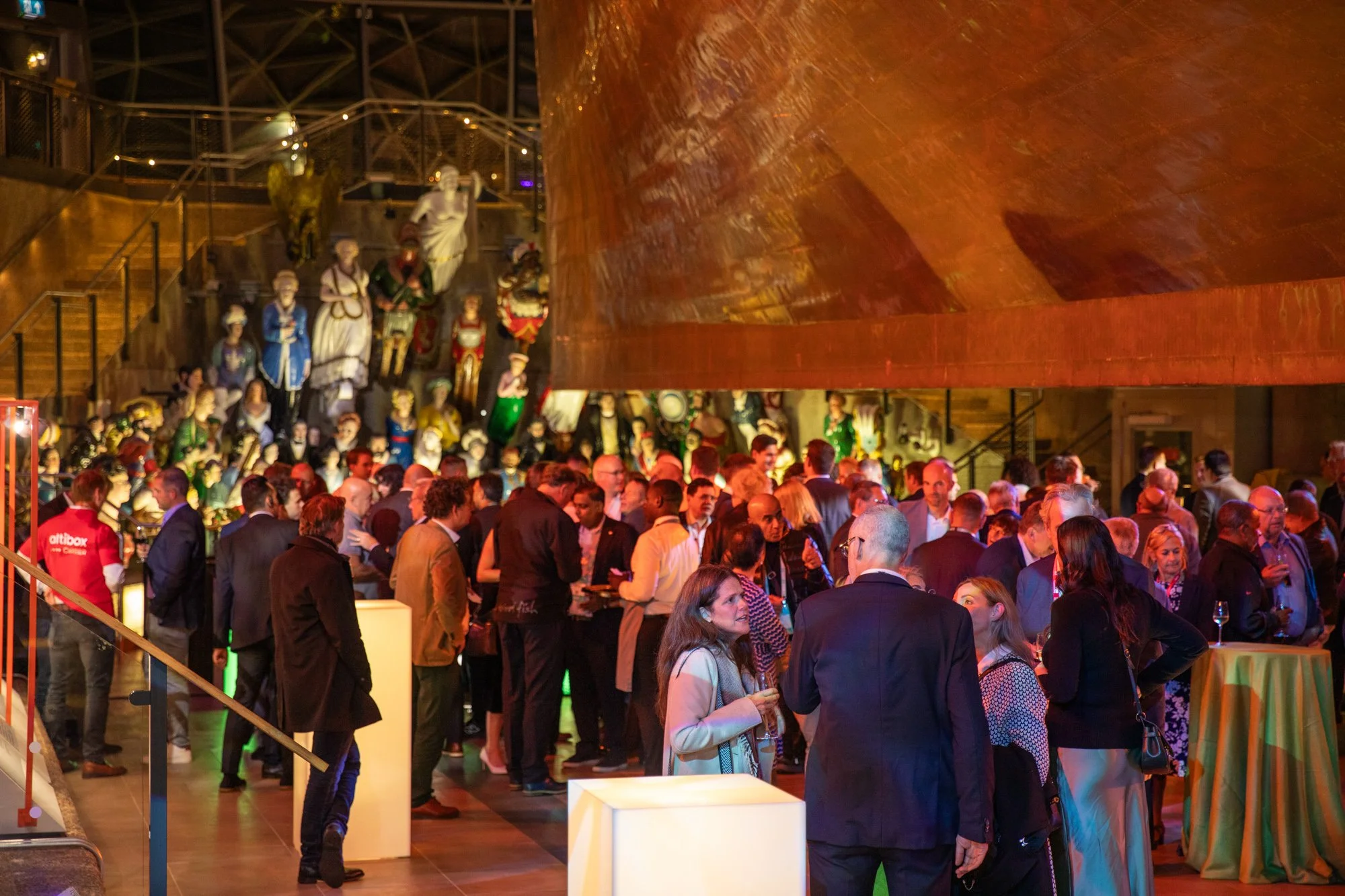 A crowded indoor event with many people socializing and enjoying drinks, with a large display of colorful figurines or sculptures in the background.