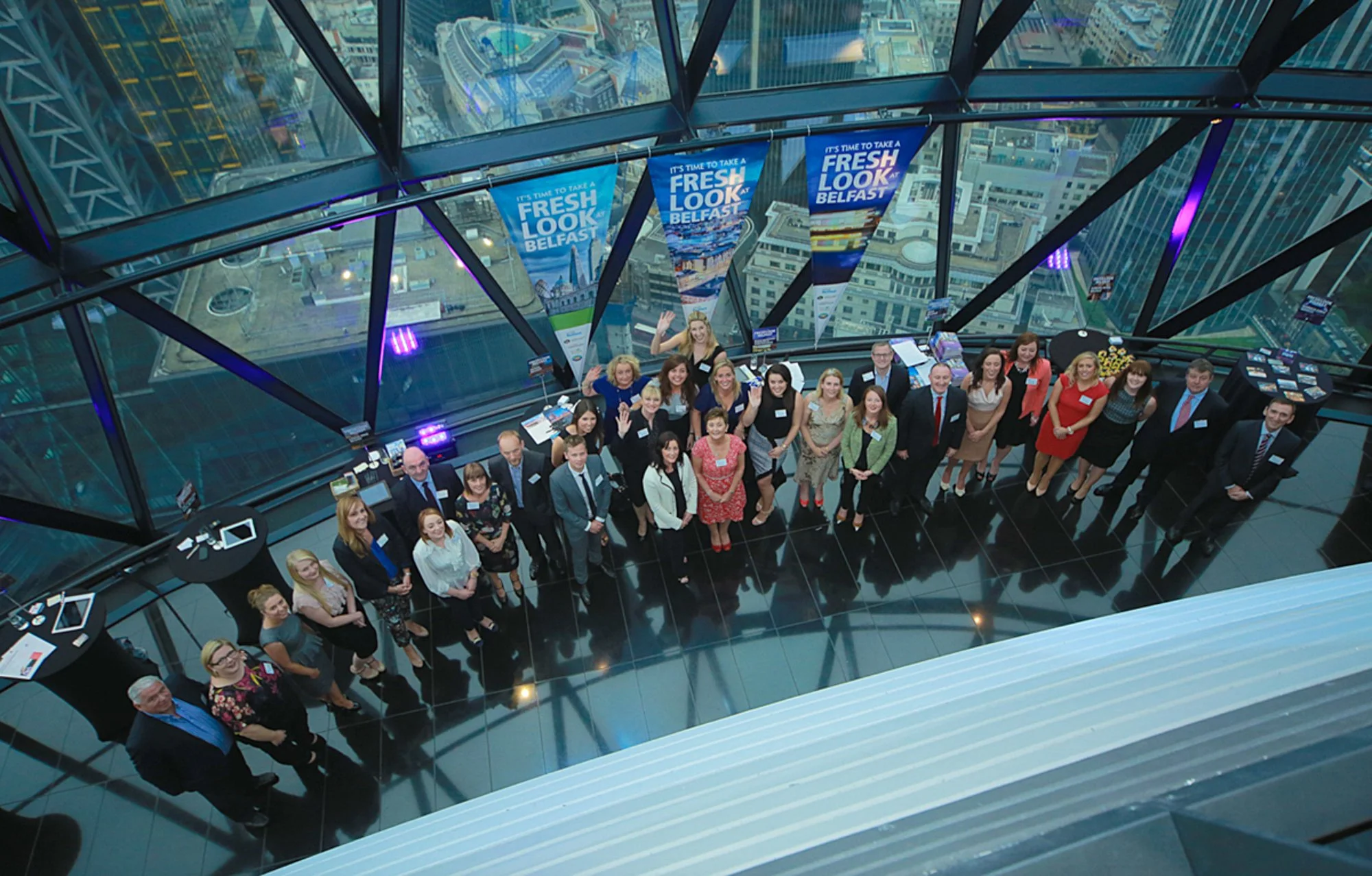 Group of people standing inside a glass tower with cityscape views, banners reading 'It's time to take a fresh look Belfast,' and informational exhibits around.