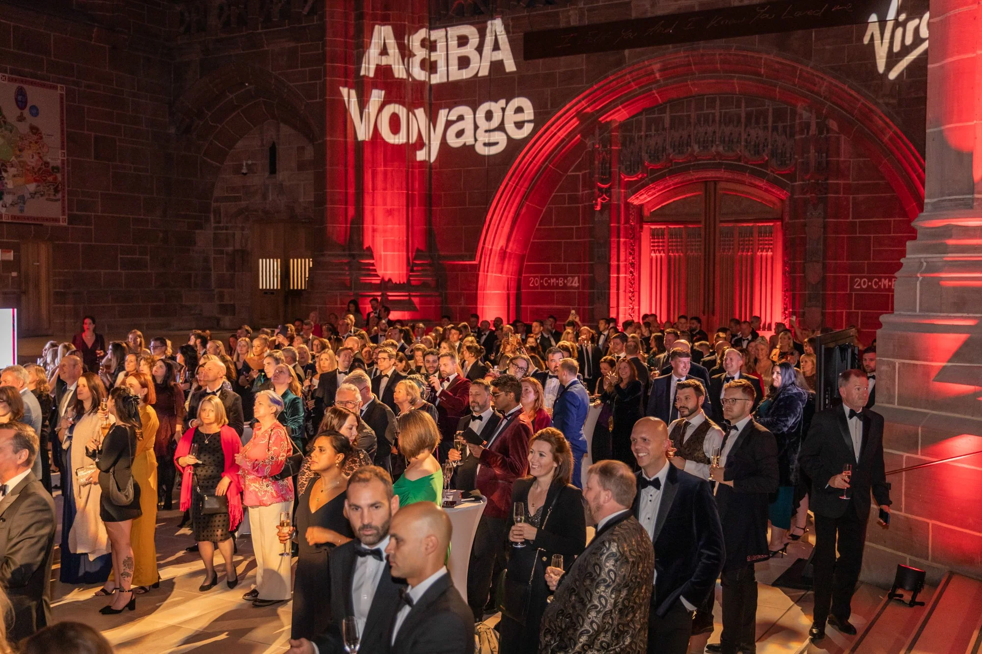 A large formal event with many people dressed in tuxedos and evening gowns, gathered in a grand hall with stone walls. The hall is illuminated with red lighting, and the words "AGBA Voyage" are projected on the wall.