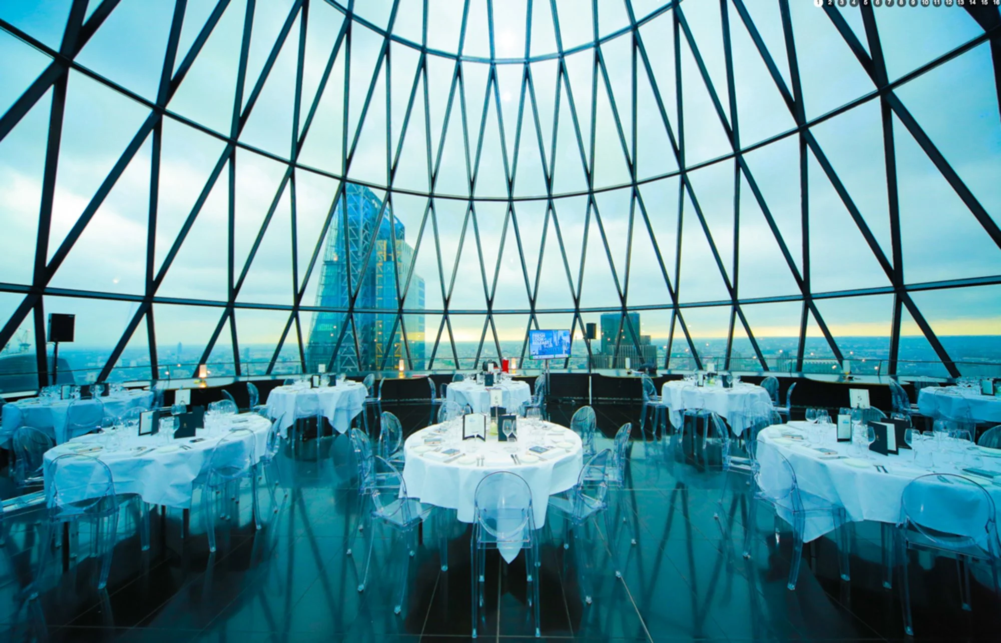 Elegant restaurant with white tablecloths and transparent chairs, set with plates and glasses, inside a modern glass building with a view of a city skyline, including a distinctive skyscraper.