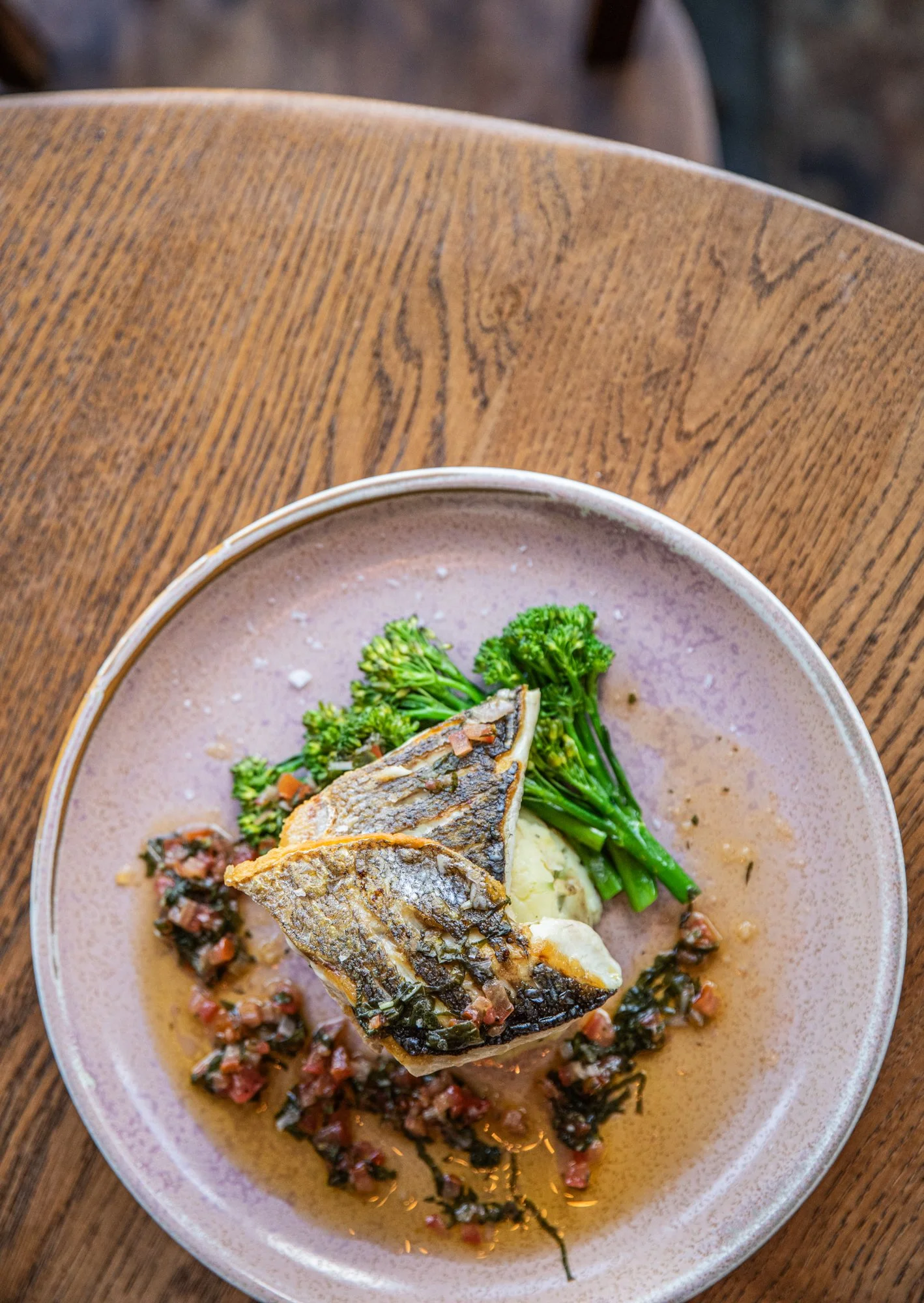 A plate with a cooked fish fillet, broccoli, mashed potatoes, and a sauce with diced vegetables, on a wooden table.