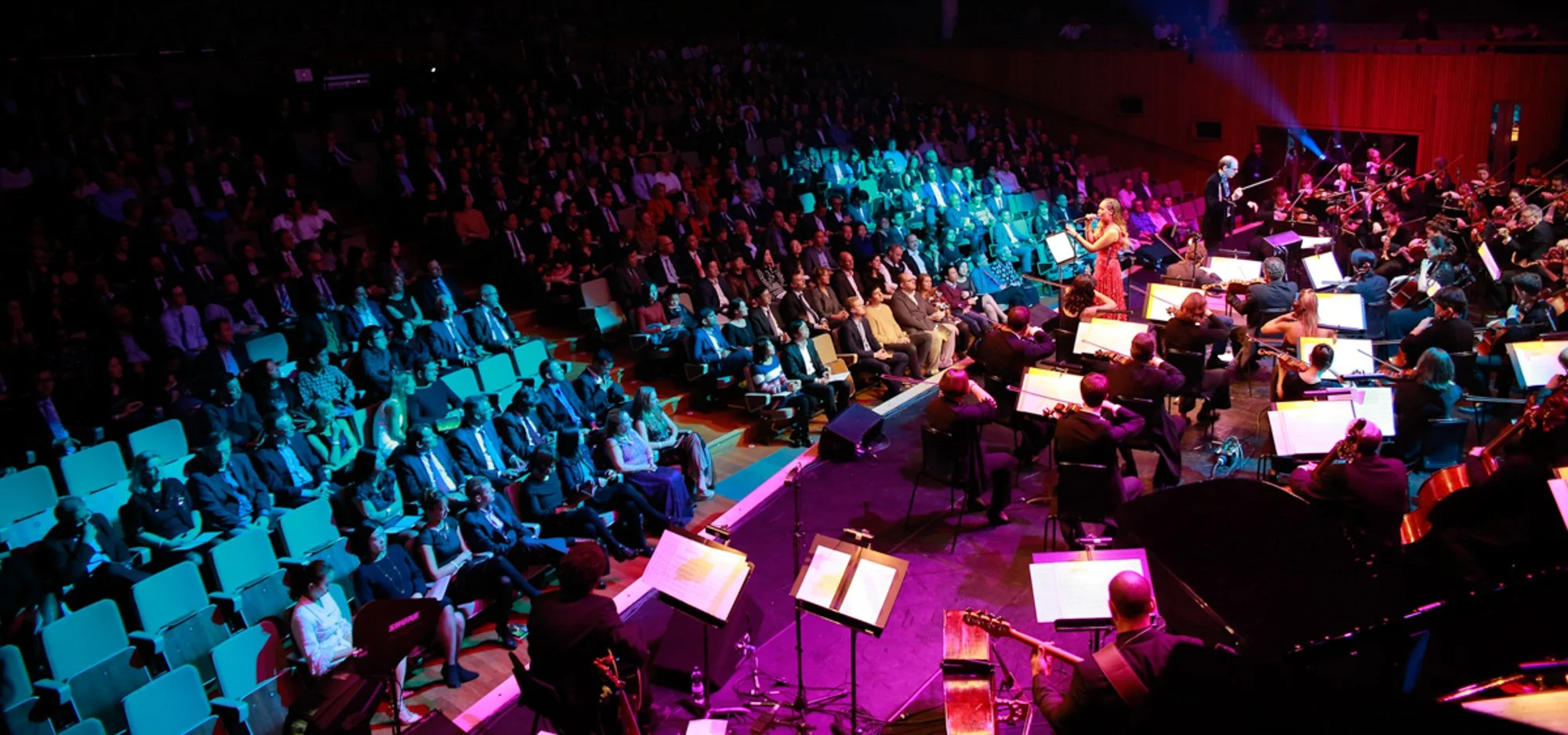 Orchestra performing on stage in a concert hall with an audience watching. The stage is lit with colorful lighting, and a female singer is at the microphones.