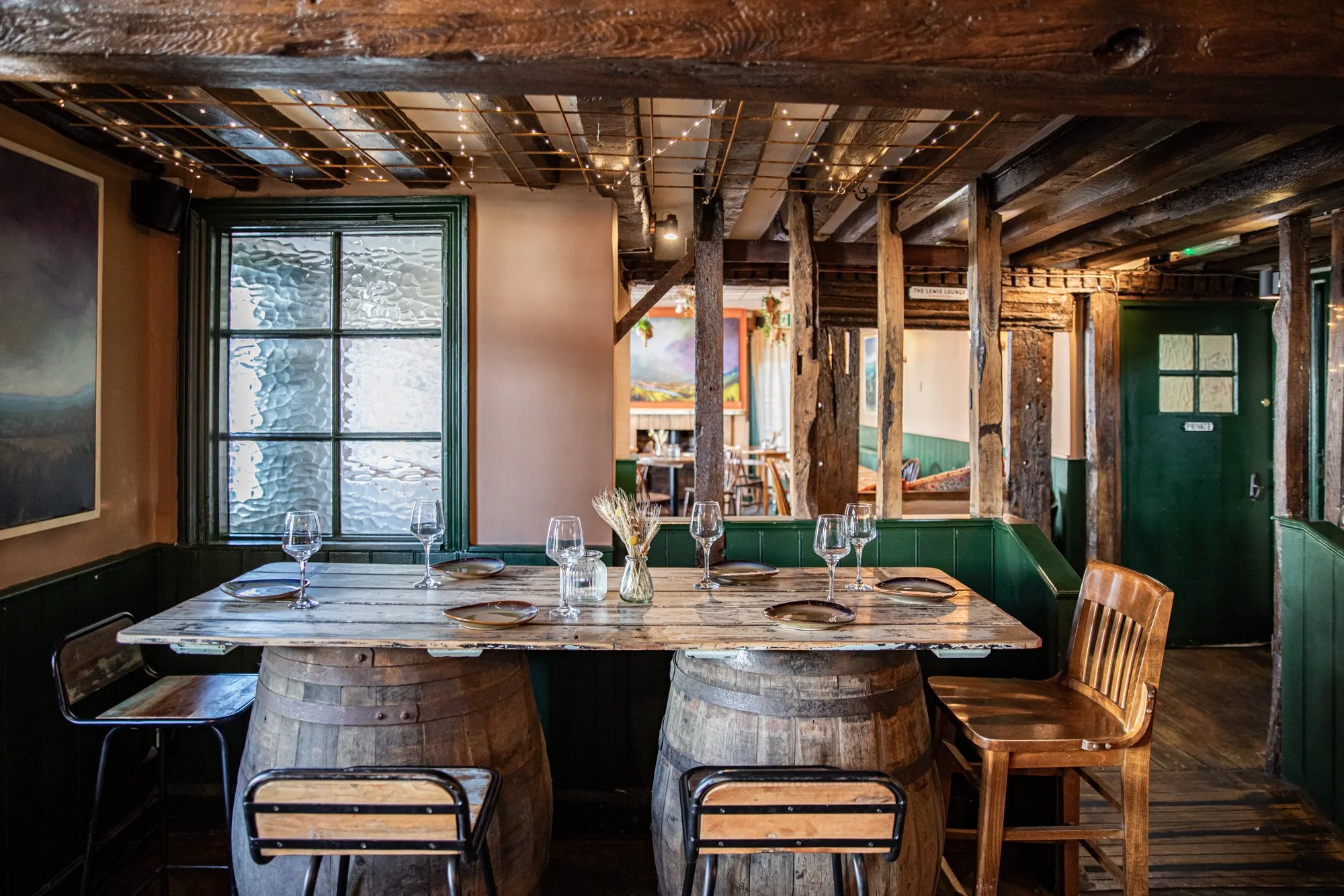 A rustic restaurant interior with a wooden table on two large barrels, set with wine glasses, plates, and a small vase with dried flowers. The room features wooden beams and string lights, with a window and artwork on the walls.