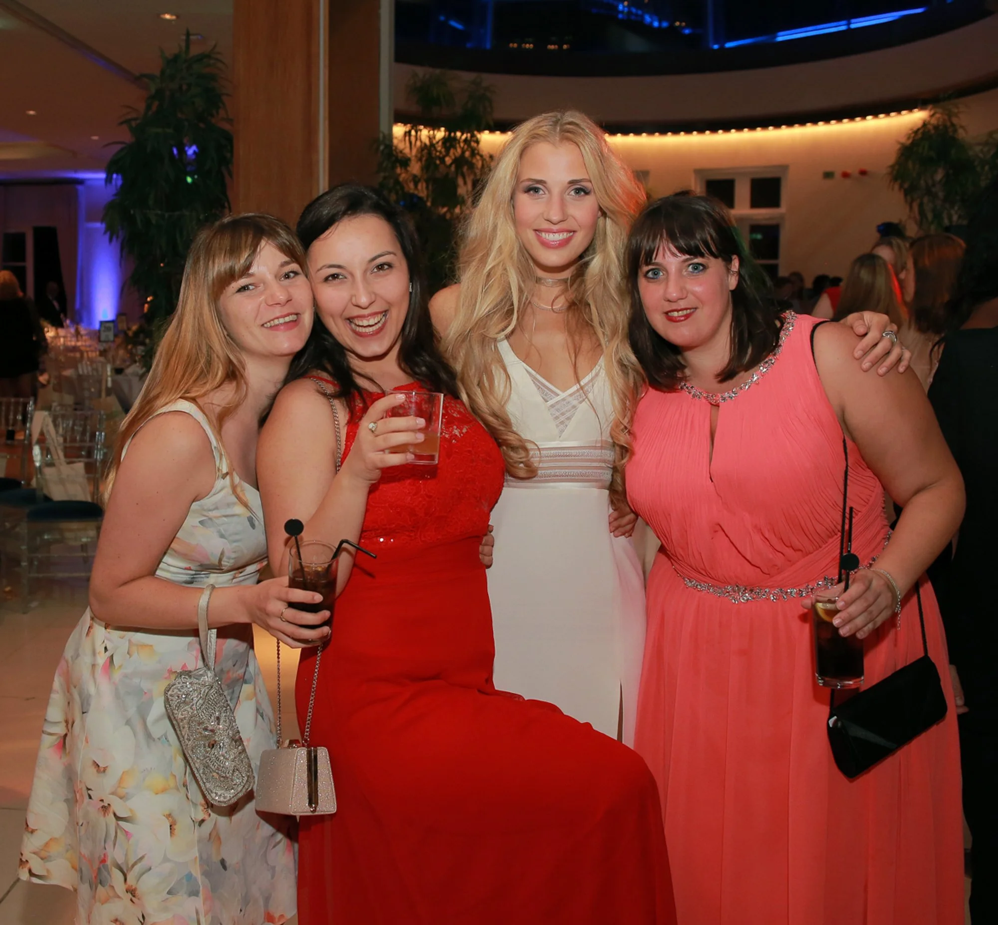 Four women at a party, smiling and holding drinks, dressed in colorful dresses, in a decorated venue with tables and plants in the background.