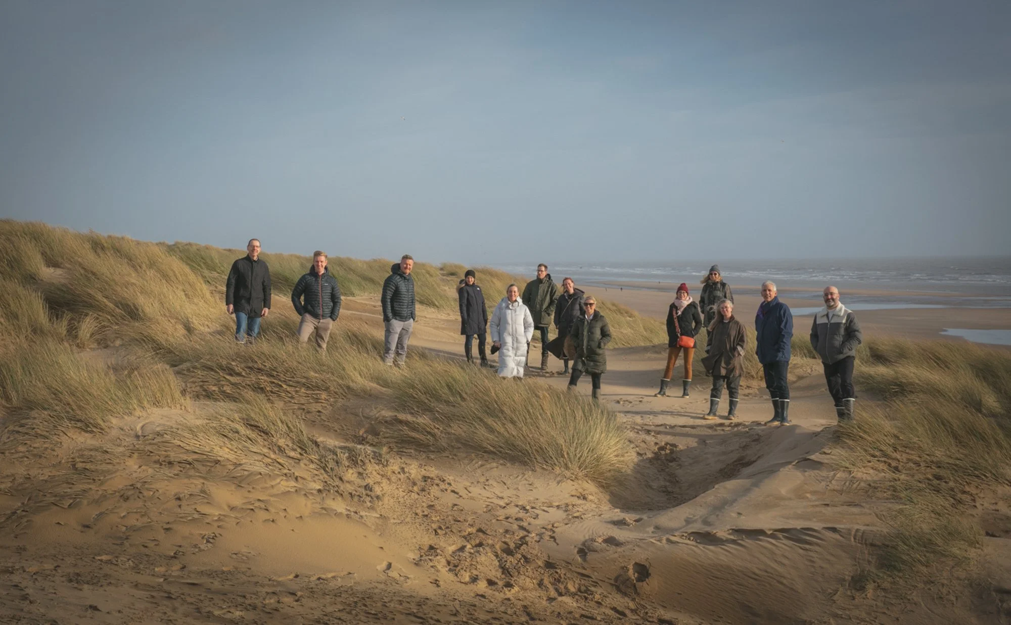 A group of fifteen people standing on sand dunes at the beach with grassy patches, ocean waves in the background, under cloudy sky.