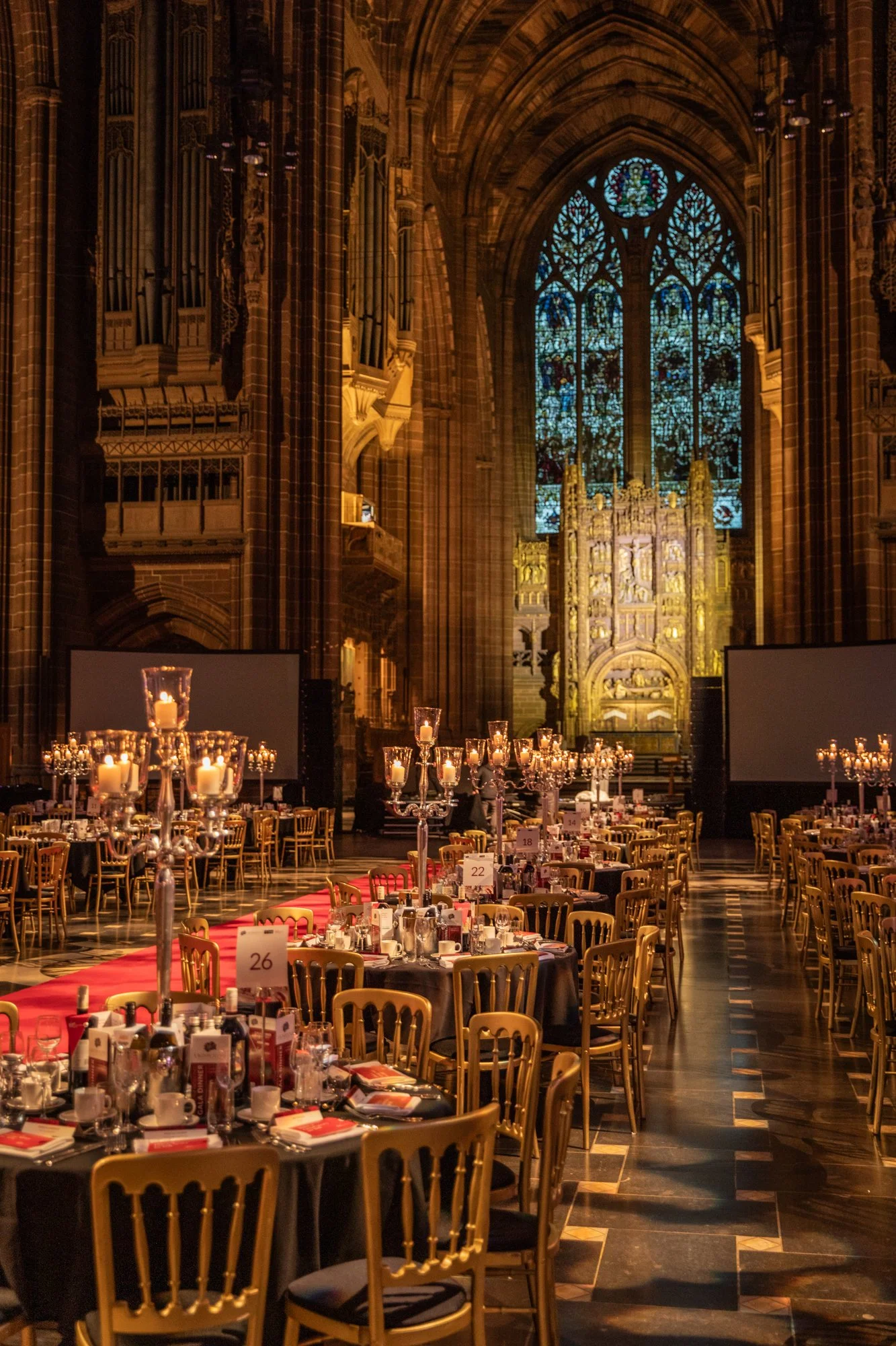 Elegant banquet hall inside a large, Gothic-style church with high vaulted ceilings and stained glass windows, decorated with numerous tall candle holders and tables set for an event.