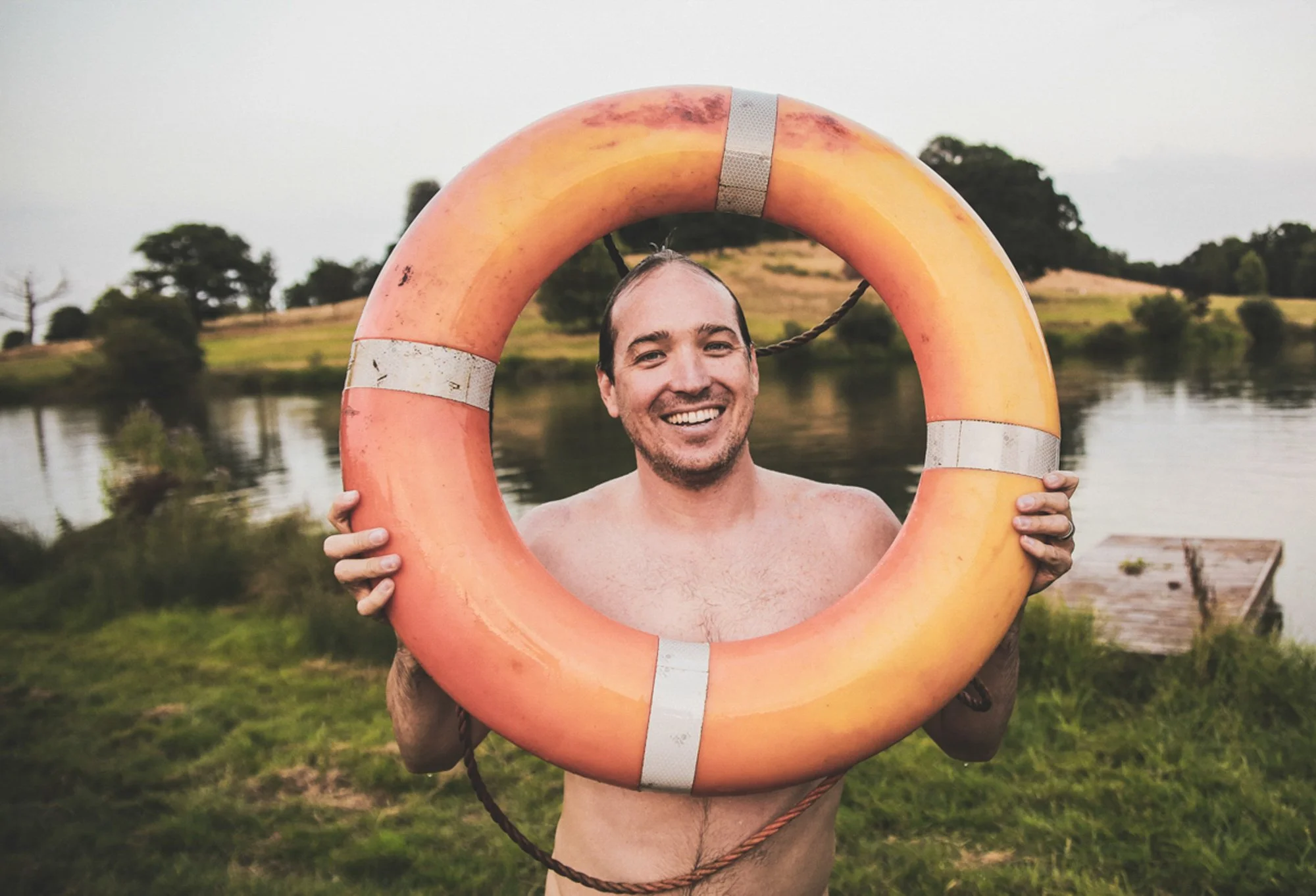 A smiling man holding a life preserver frame around his face near a lake with grassy hills and trees in the background.