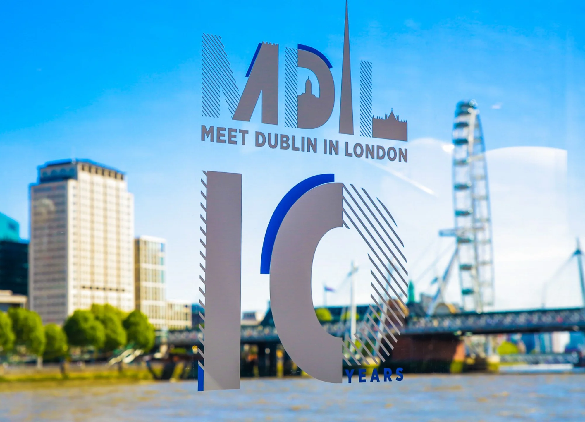 Reflection of a sign on a window with a cityscape in the background, including a ferris wheel and a bridge. The sign reads "APC Meet Dublin in London 10 Years"